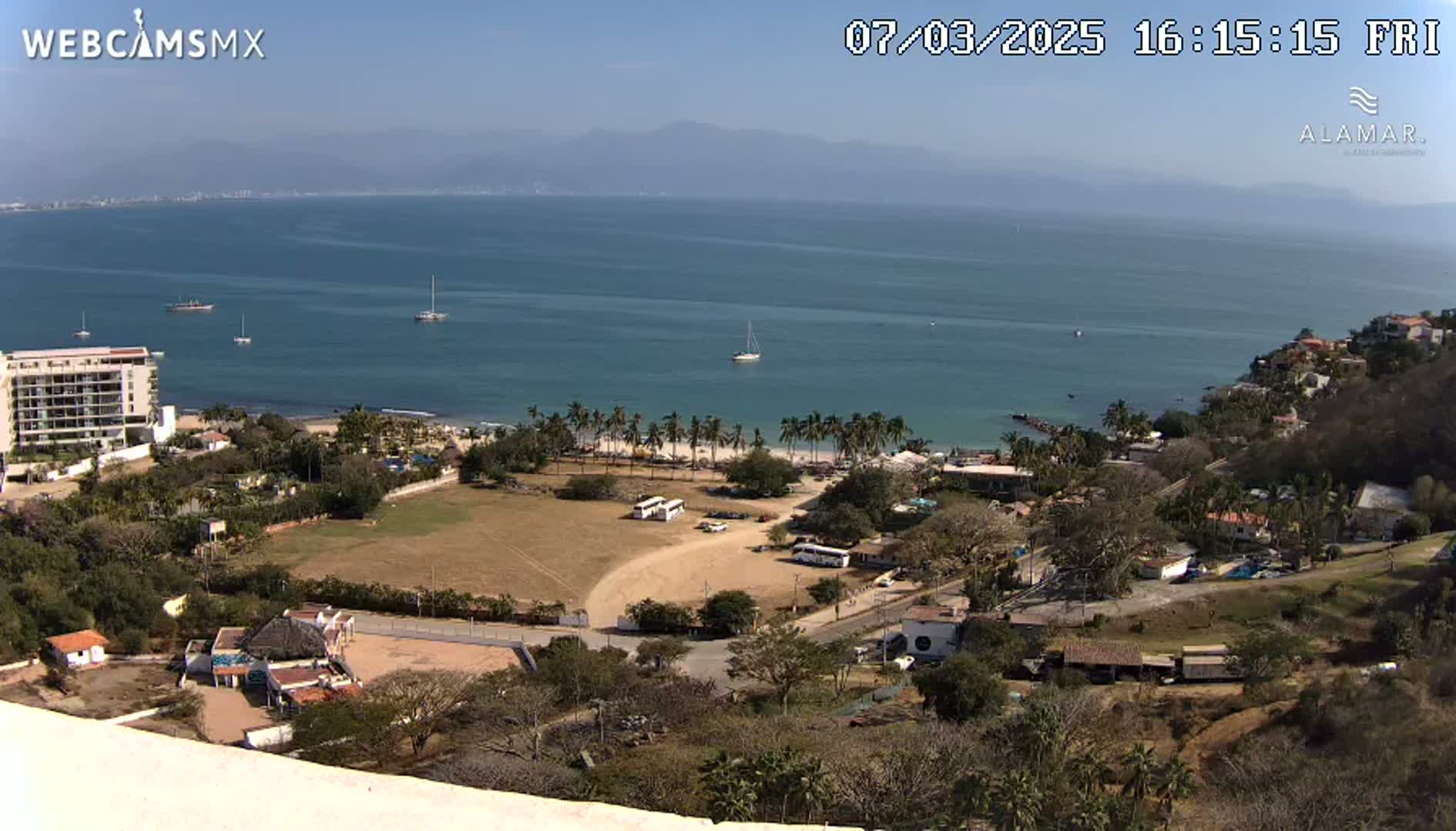A coastal town overlooking a calm bay on a sunny day, with several boats visible in the water and palm trees lining the beach.