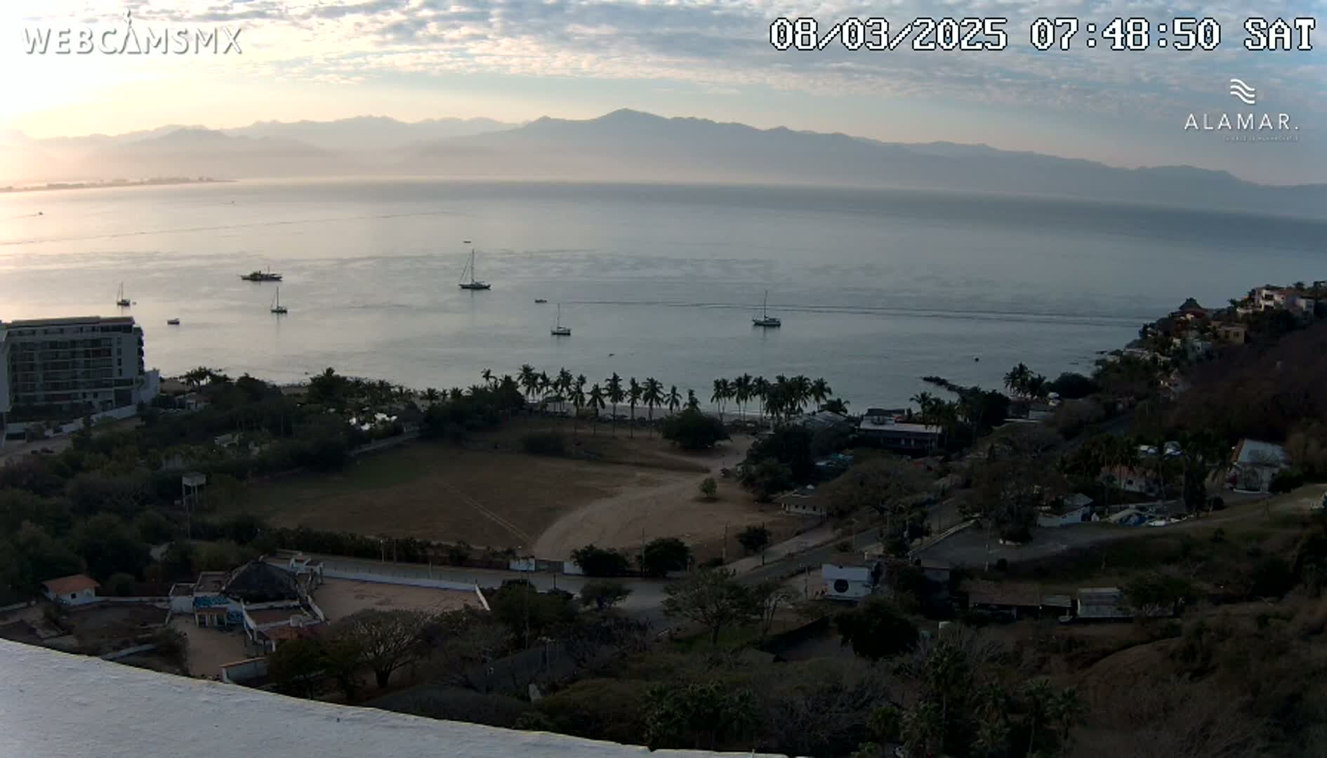 A high-angle, long shot shows a calm bay with several boats, a coastline with buildings and vegetation, and mountains in the distance under a partly cloudy, dawn sky.