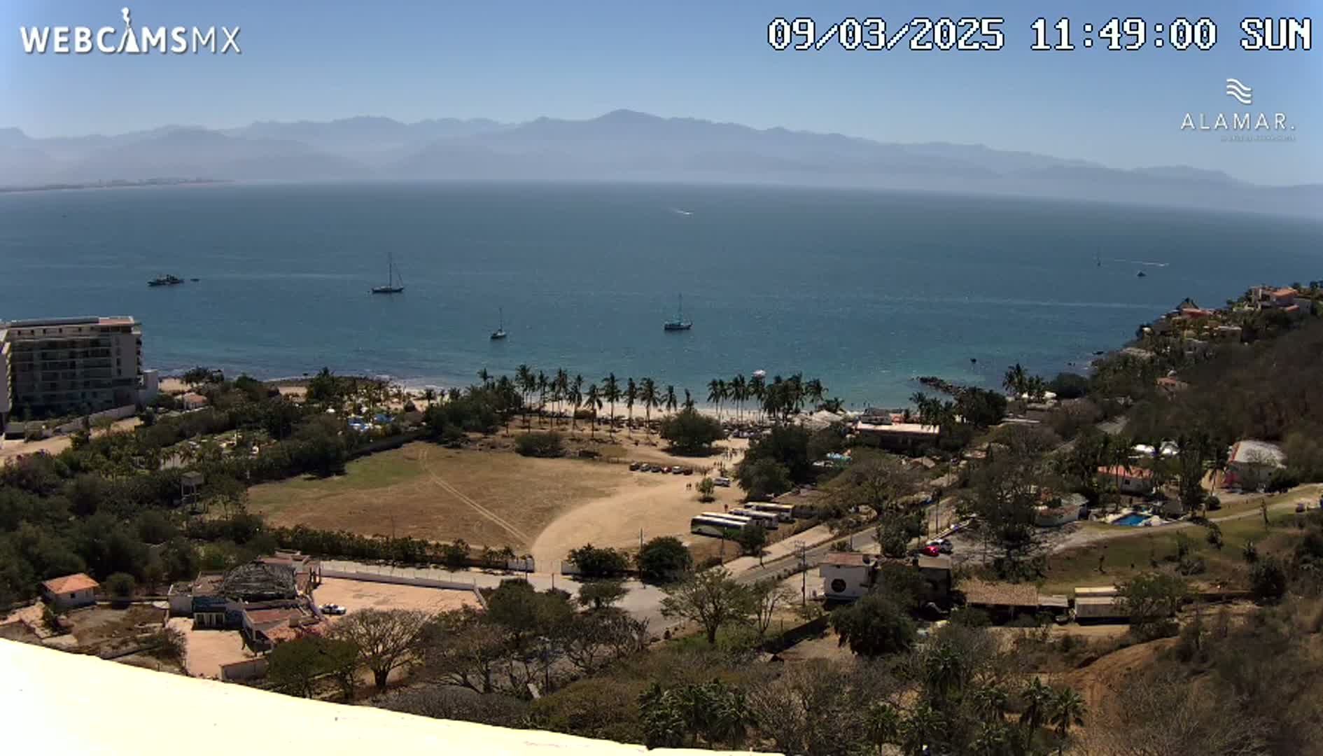 A sunny coastal scene shows a calm ocean with several boats, a sandy beach lined with palm trees, and a hillside town with buildings and vegetation, all under a clear blue sky.