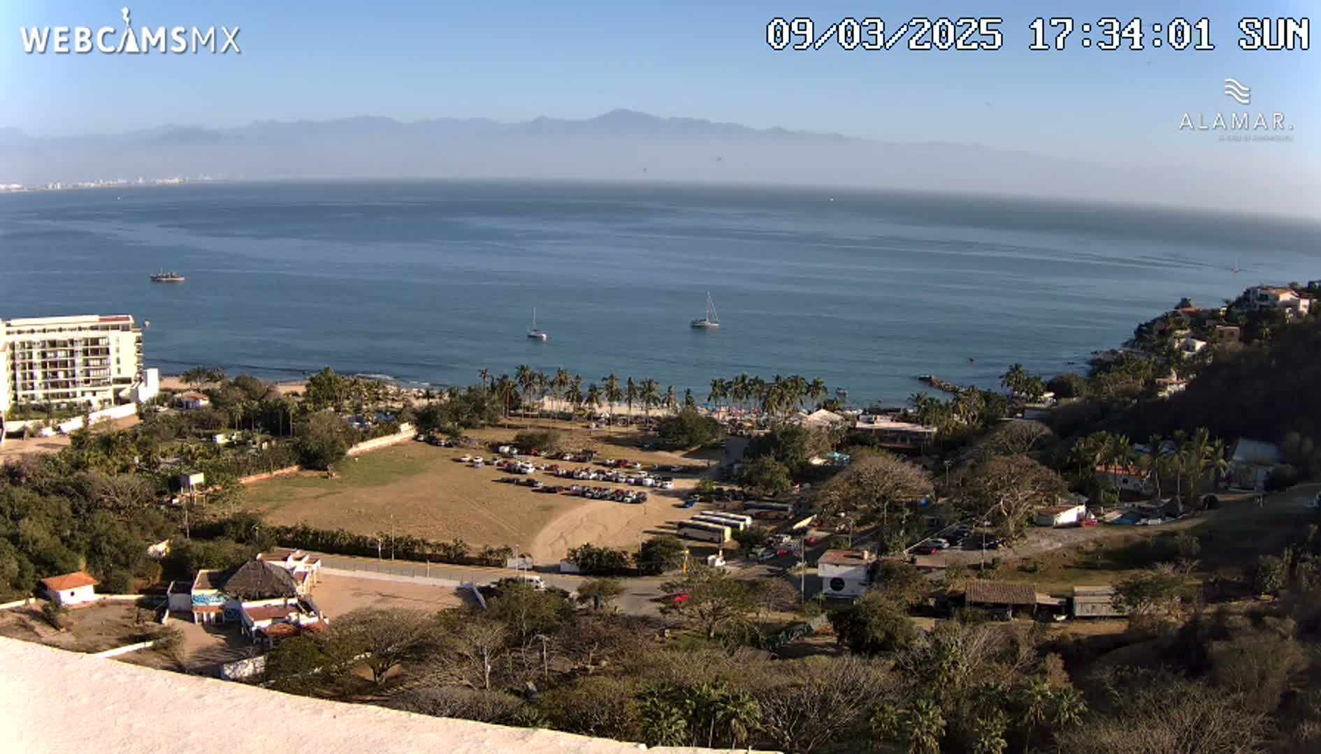 A high-angle, sunny view of a coastline shows a calm ocean with several boats, a beach with palm trees, buildings, and a parking lot, all situated beneath a clear sky and distant mountains.