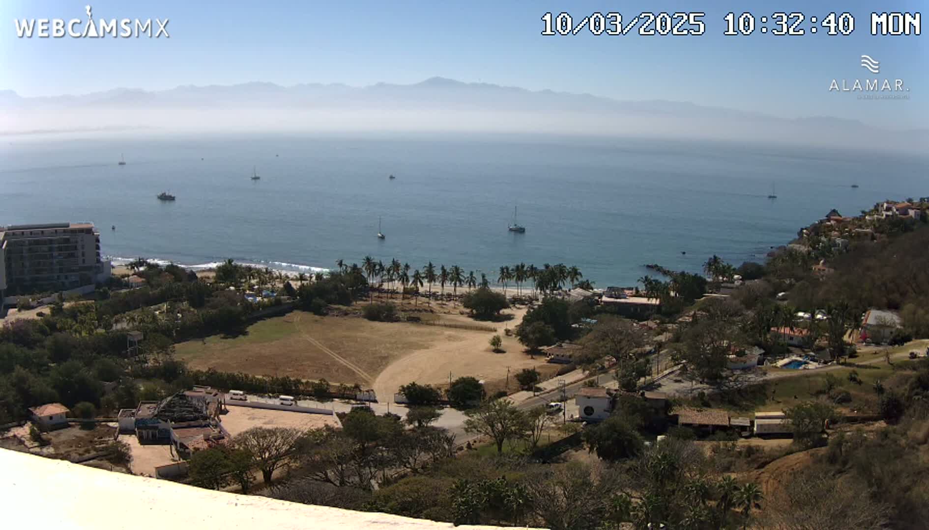 A high-angle, long shot of a coastline on a clear, sunny day, showing a beach with palm trees, buildings, and a calm ocean with several boats visible in the distance, and mountains in the far background.