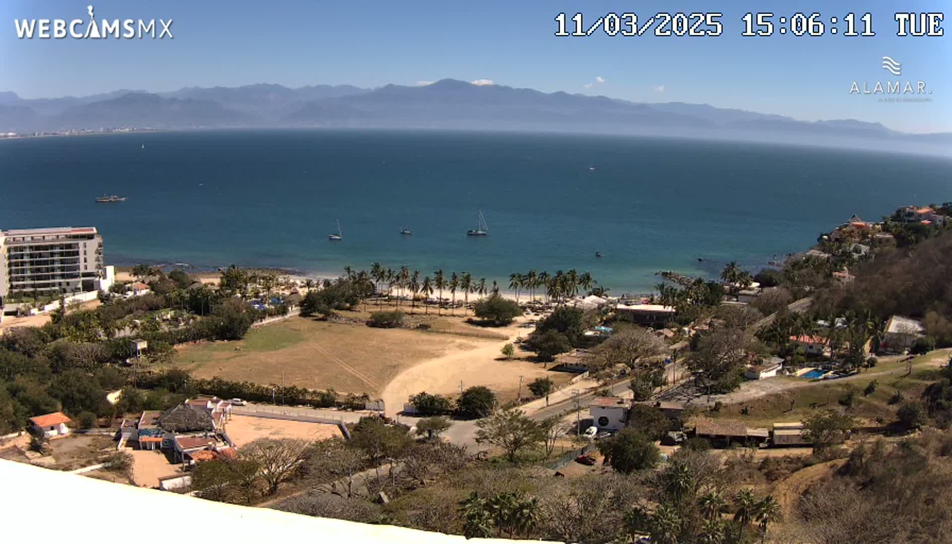 A high-angle view shows a clear, sunny day overlooking a coastline with a calm ocean, sandy beach lined with palm trees, buildings, and dry, brown land.