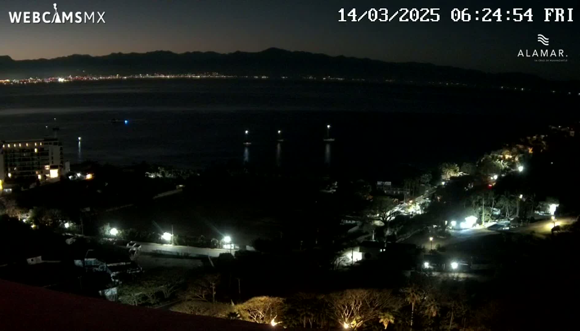 A nighttime coastal scene shows a dark body of water with a distant city skyline illuminated, a resort area with lights visible on the land, and dark mountains in the background under a clear night sky.