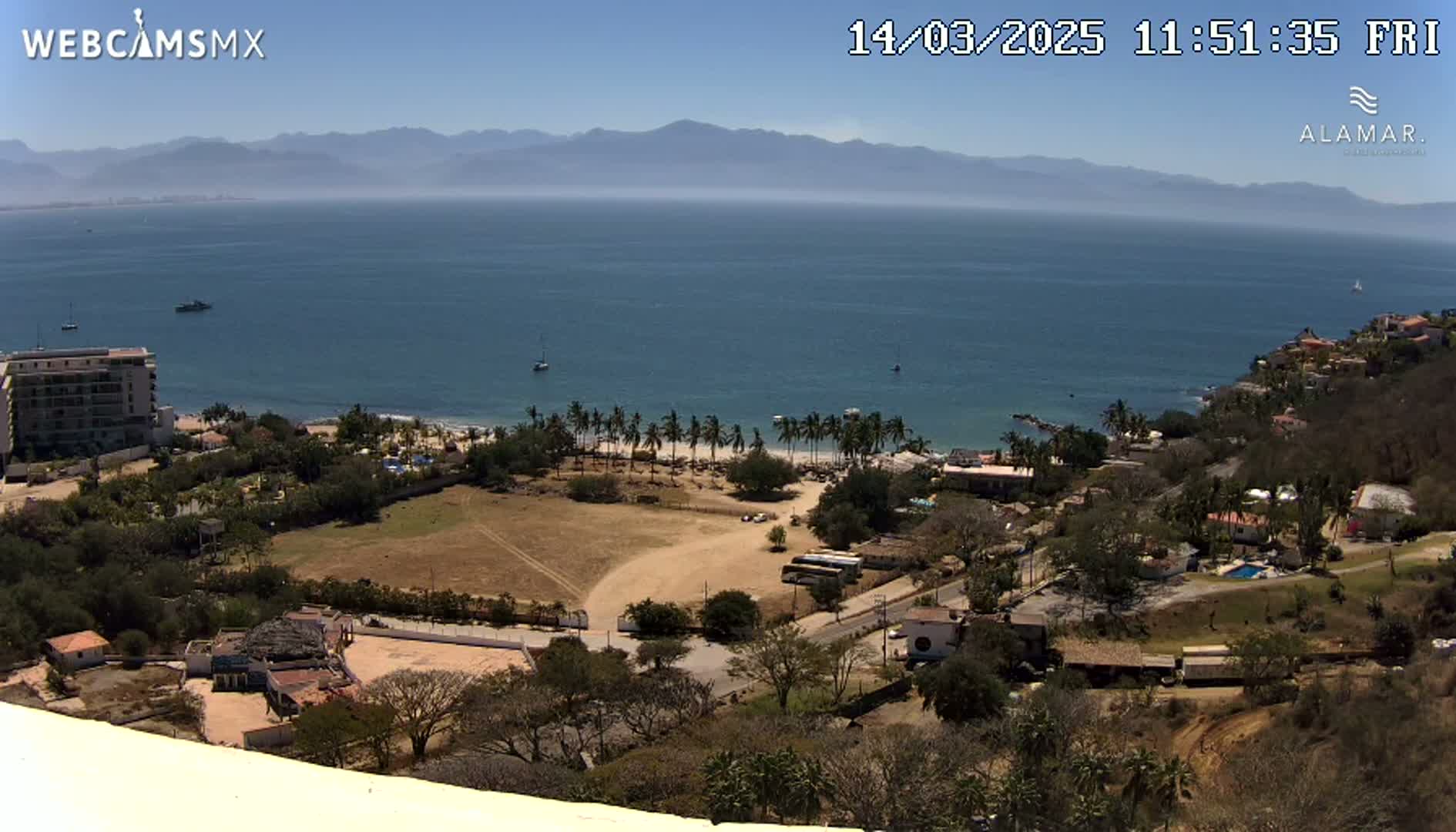 A high-angle view of a coastal town on a clear, sunny day, showing a sandy beach lined with palm trees, a calm ocean, and distant mountains.