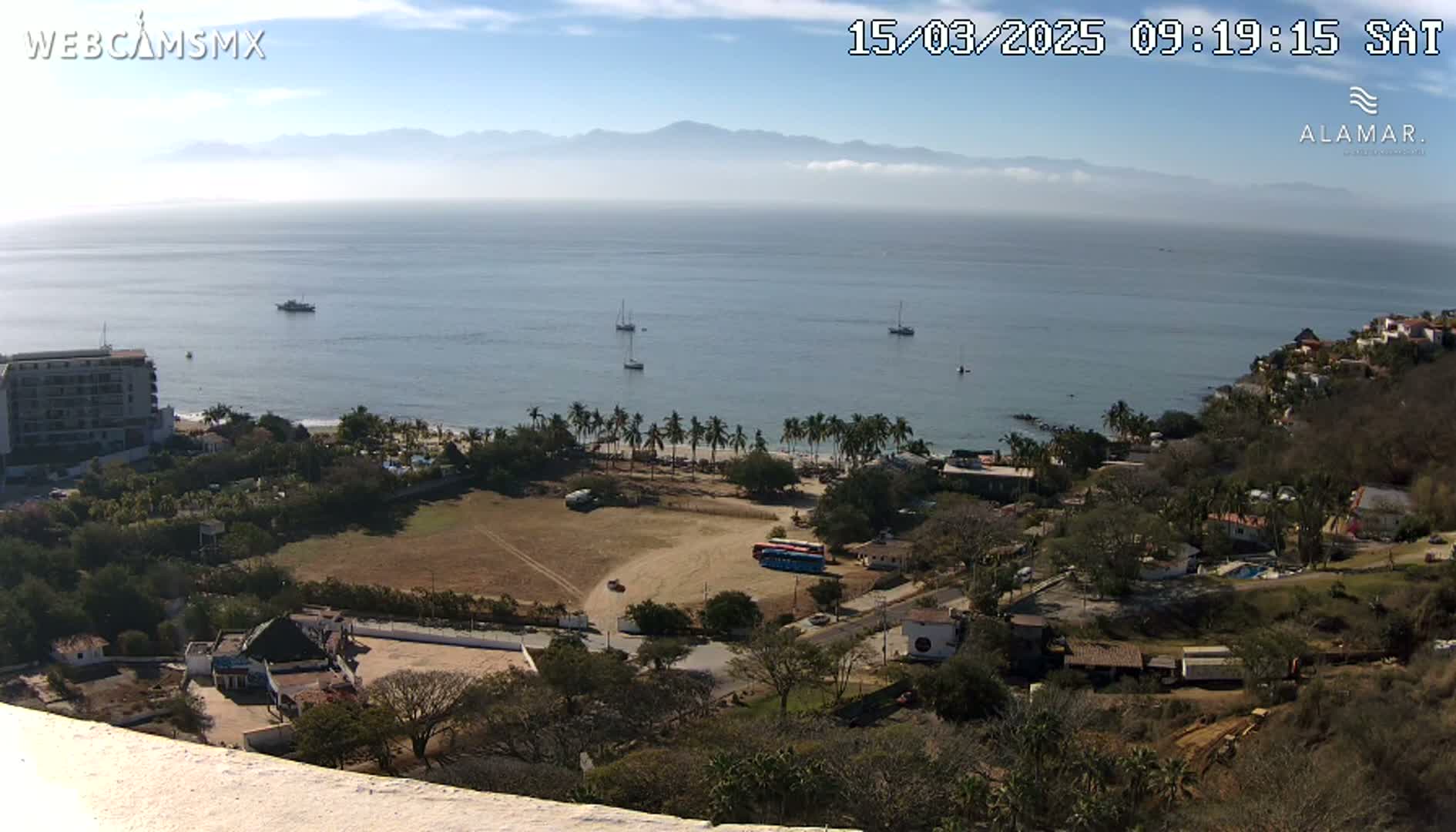 A high-angle, long shot on a sunny day shows a coastal town with a sandy beach, palm trees, buildings, and a few boats in the calm ocean with hazy mountains in the background.