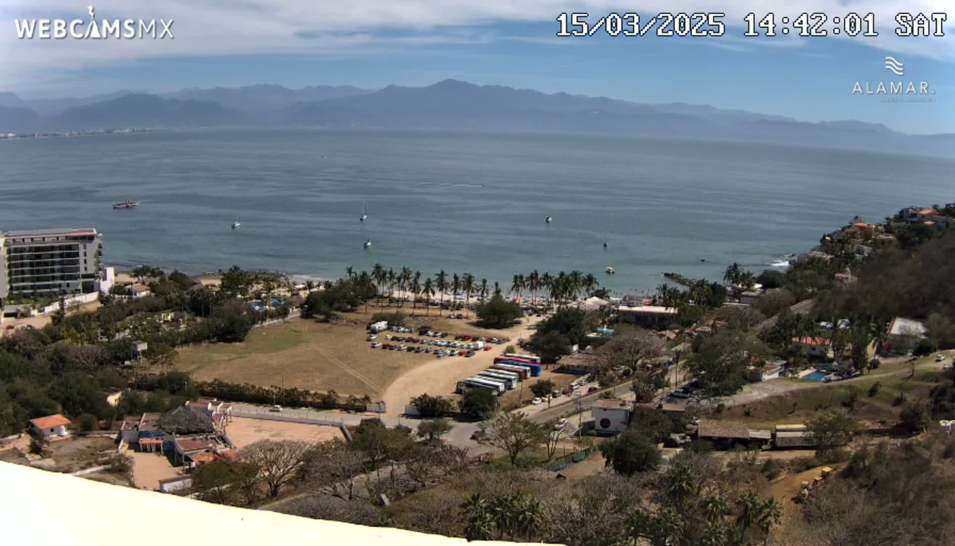 A high-angle, sunny view of a coastal town with a sandy beach, palm trees, a parking lot filled with cars and buses, and a calm ocean with distant mountains under a partly cloudy sky.