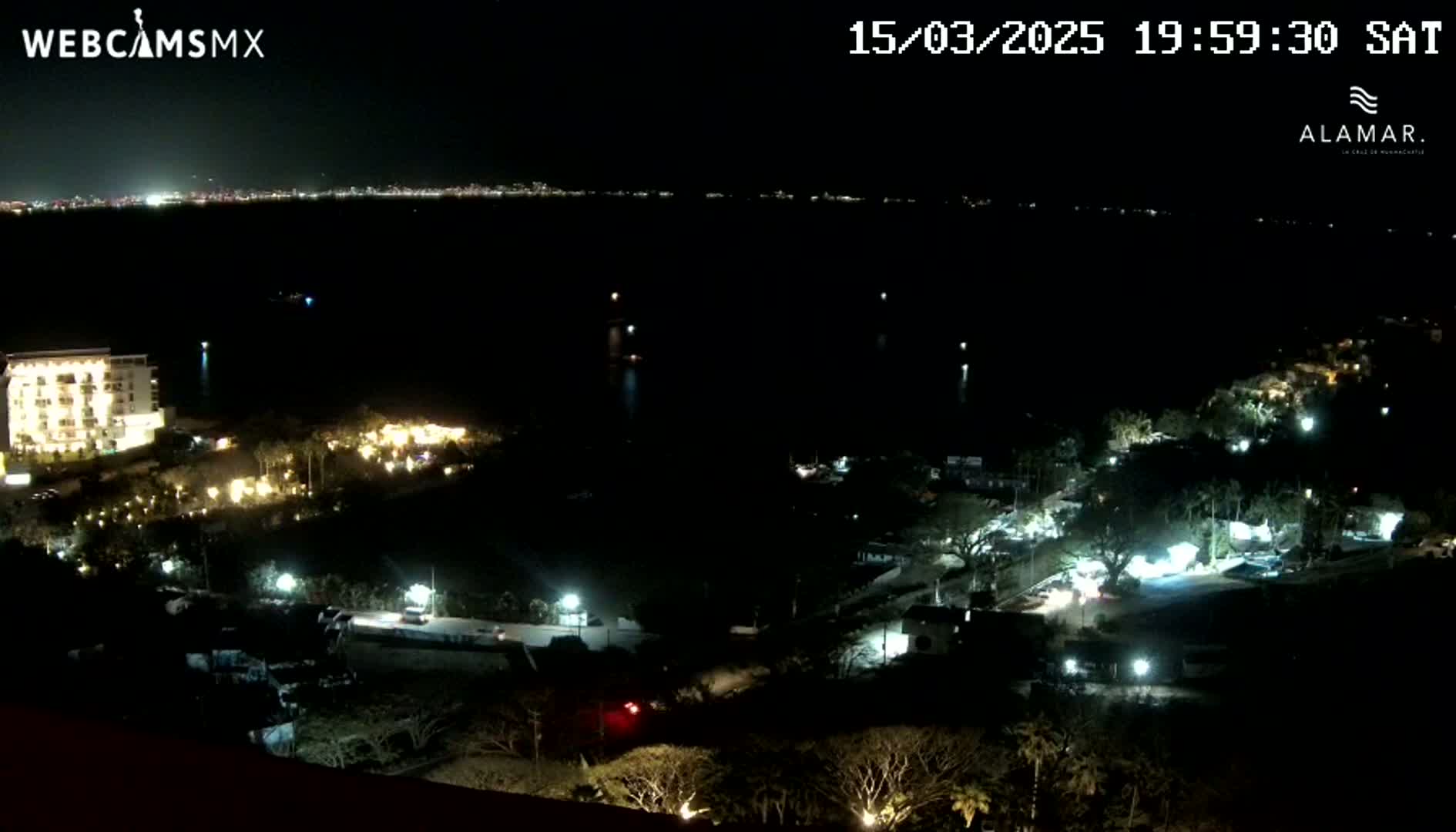 A nighttime view of a coastal area shows a city skyline across a dark body of water, with illuminated buildings and landscaping closer to the camera.