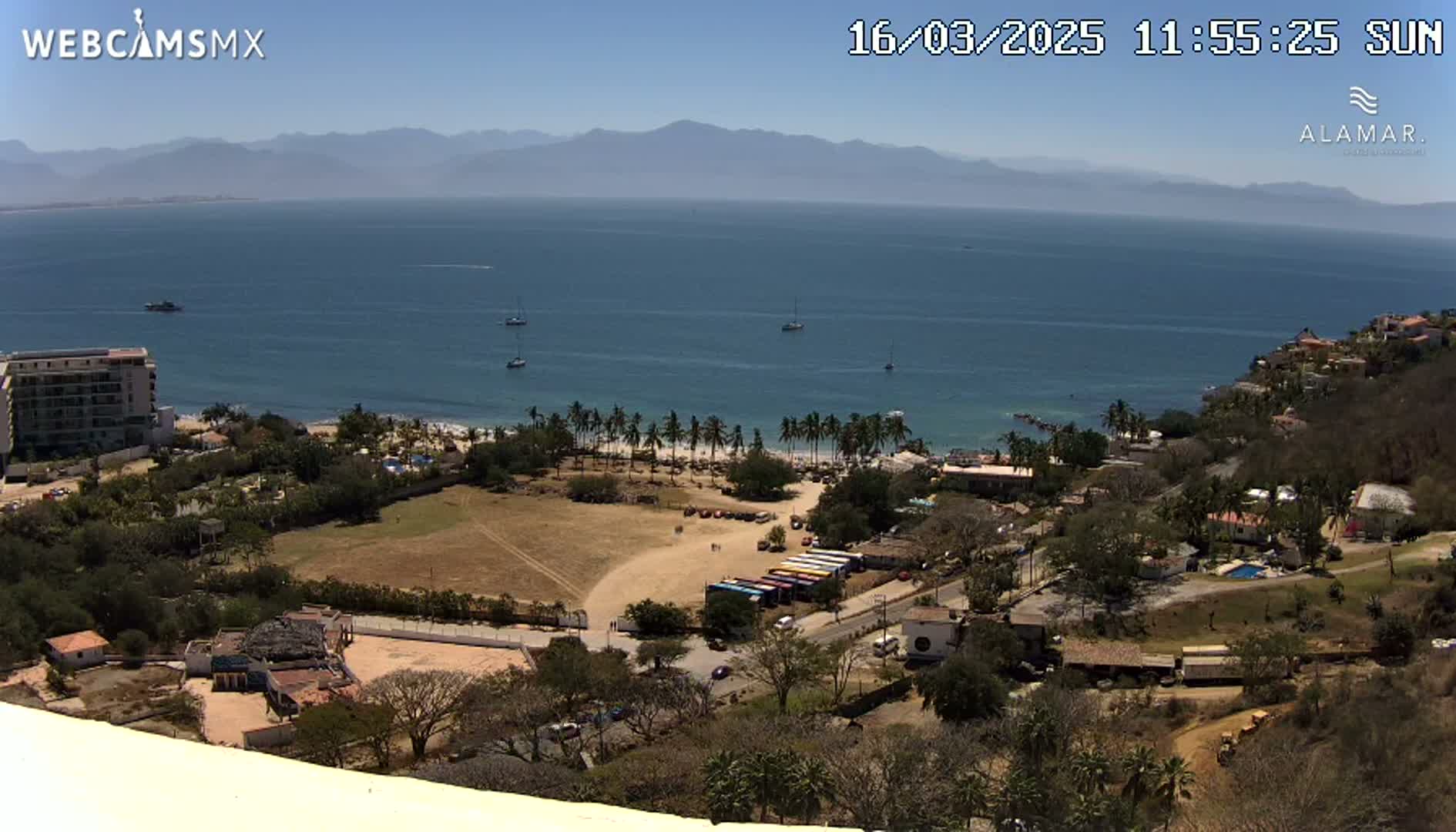 A high-angle, sunny view shows a coastal town with a sandy beach, palm trees, and several boats in the ocean, all set against a backdrop of distant mountains.