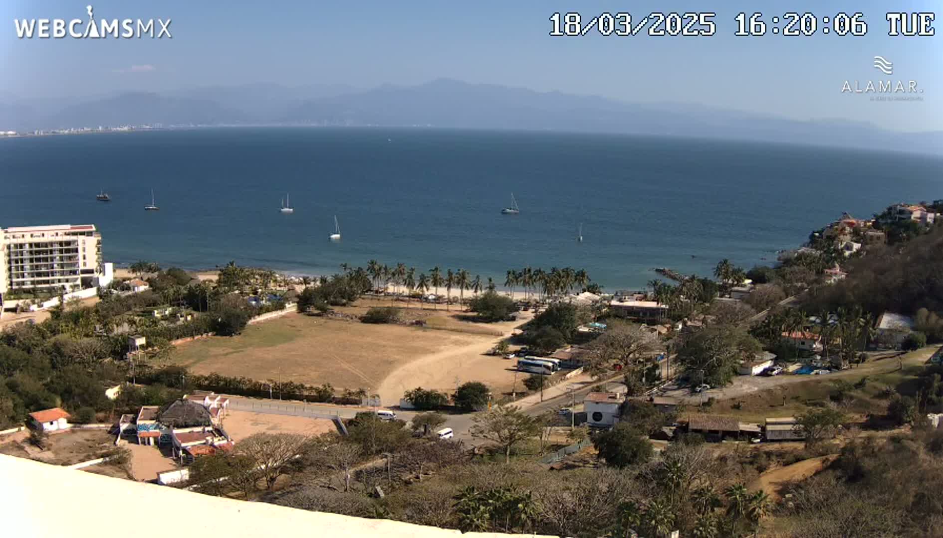 An elevated view shows a coastal town on a sunny day, with a calm ocean in the background and several sailboats visible.