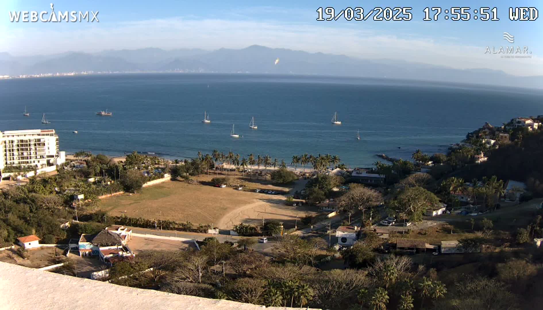 A high-angle view of a coastal town on a sunny day, with several sailboats visible in a calm bay, mountains in the distance, and a sandy beach lined with palm trees.