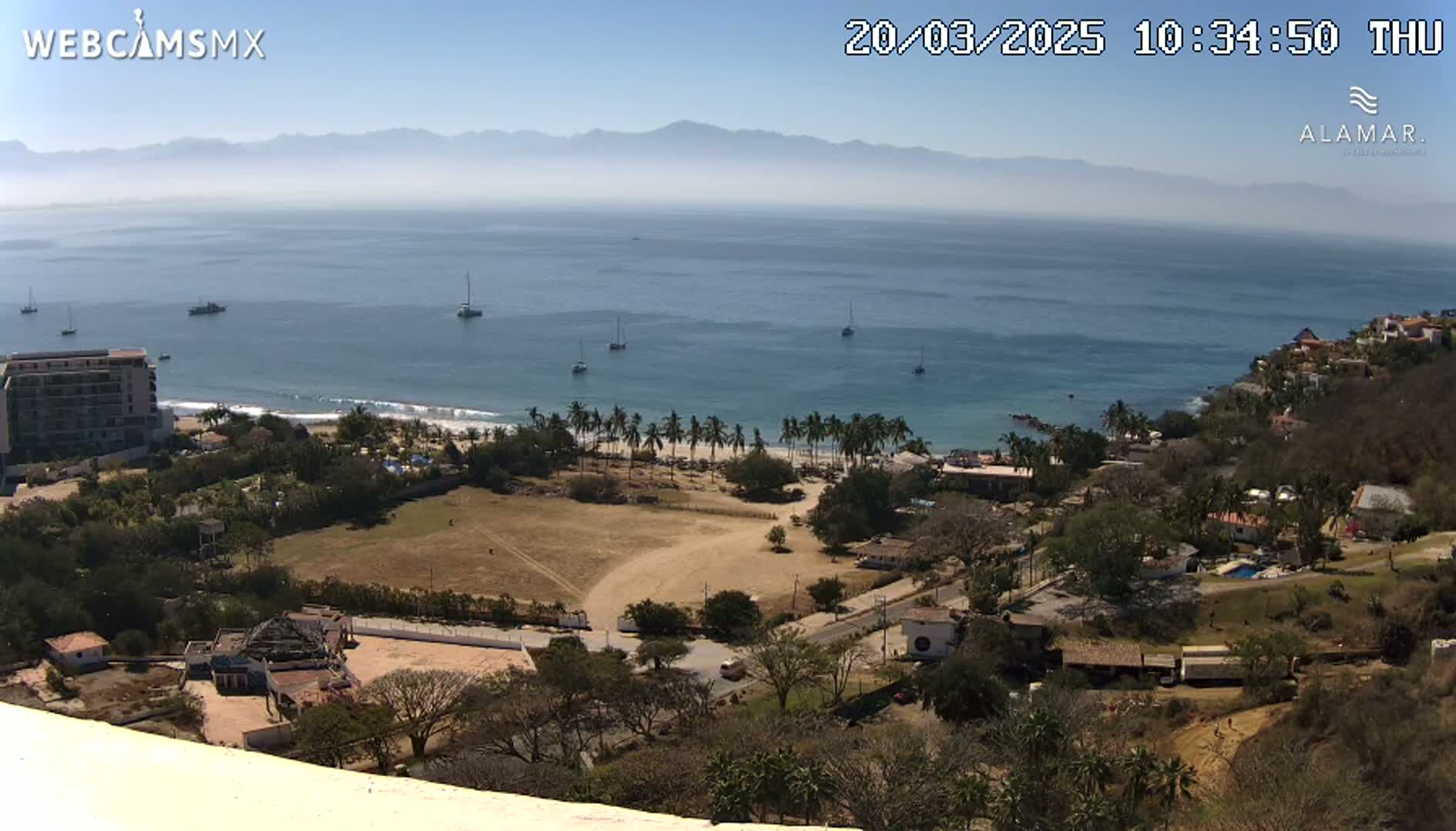 An elevated view shows a coastal town on a sunny day, with a calm ocean, a sandy beach lined with palm trees, several sailboats at sea, and distant mountains under a clear sky.