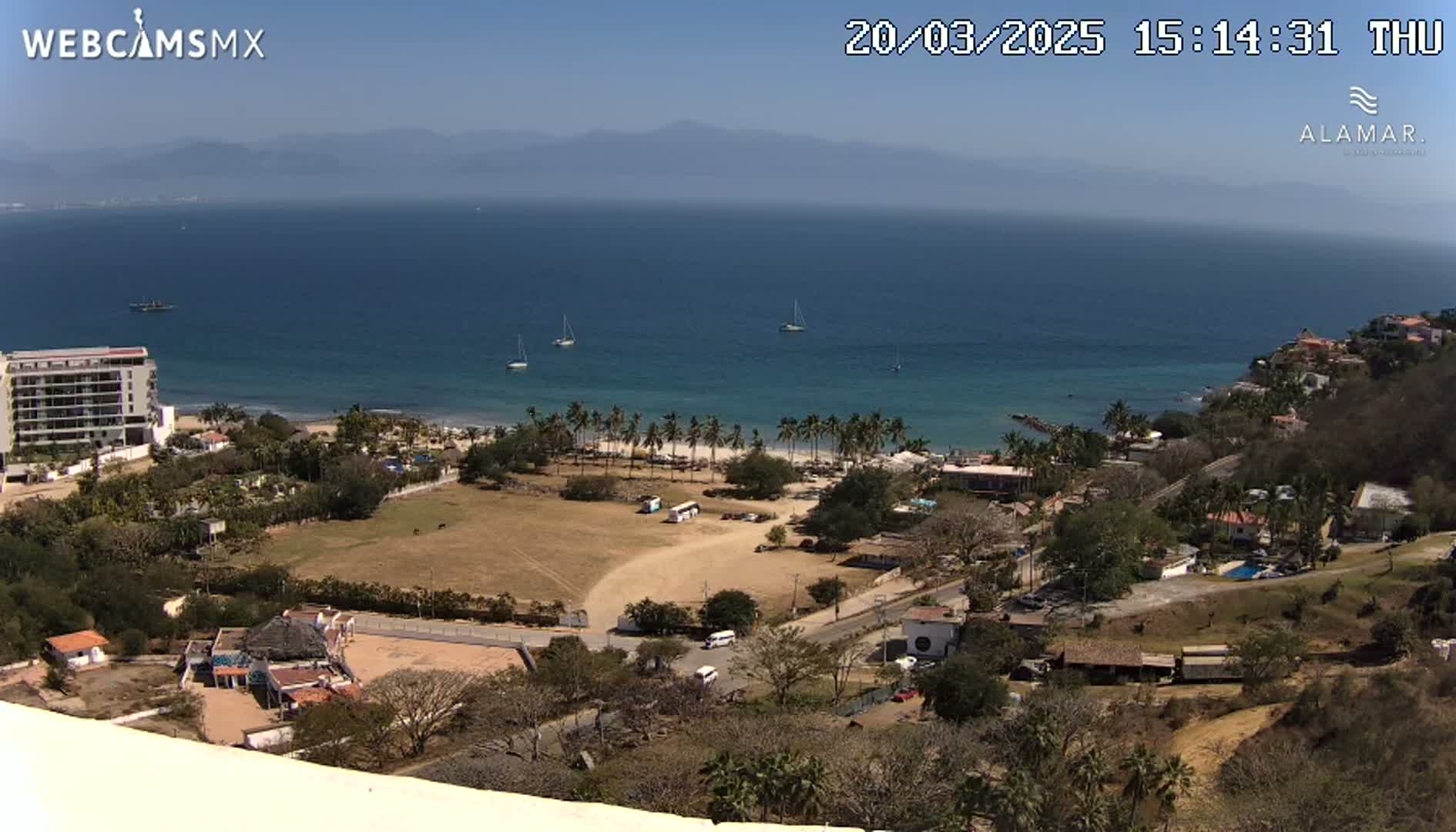A high-angle view on a sunny day shows a coastline with a sandy beach lined with palm trees, a calm ocean with several boats, and buildings nestled among dry, scrubby vegetation.