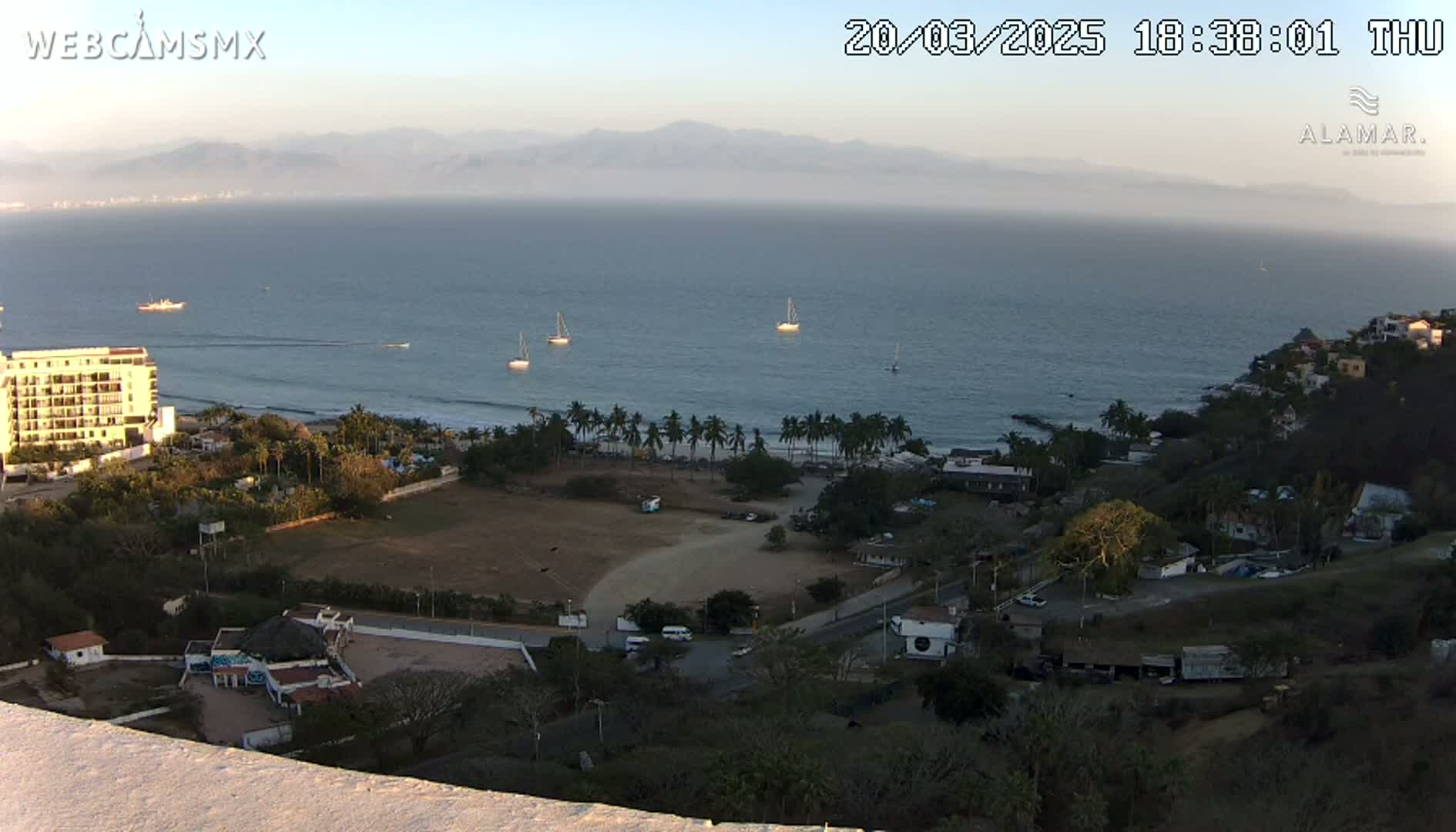 An elevated view of a coastal town at sunset, showing a calm ocean with several sailboats, a beach lined with palm trees, buildings, and a large empty field.