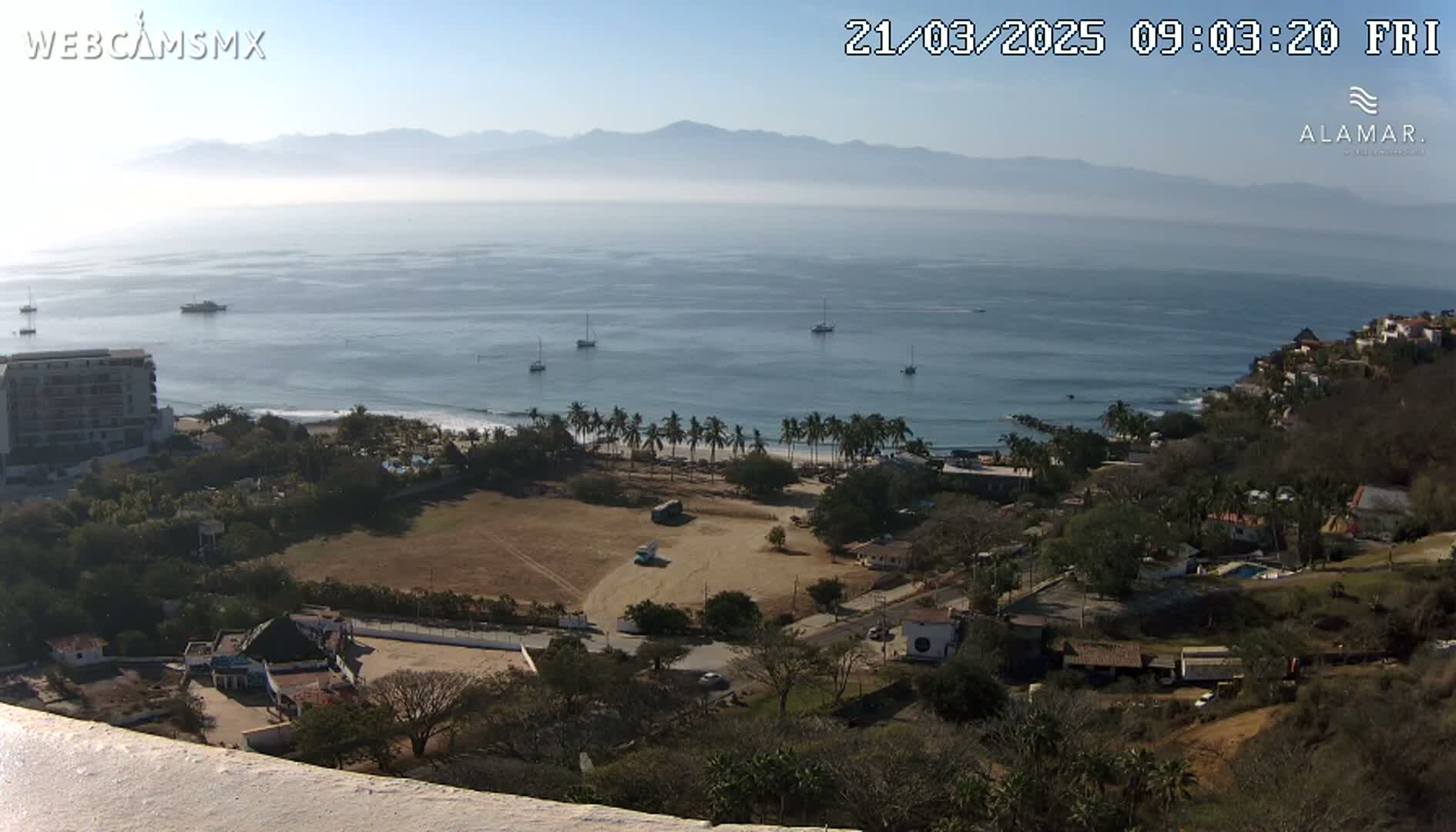 A coastal scene on a hazy, sunny day shows a sandy beach lined with palm trees, buildings, and several boats on calm ocean waters, with distant mountains in the background.