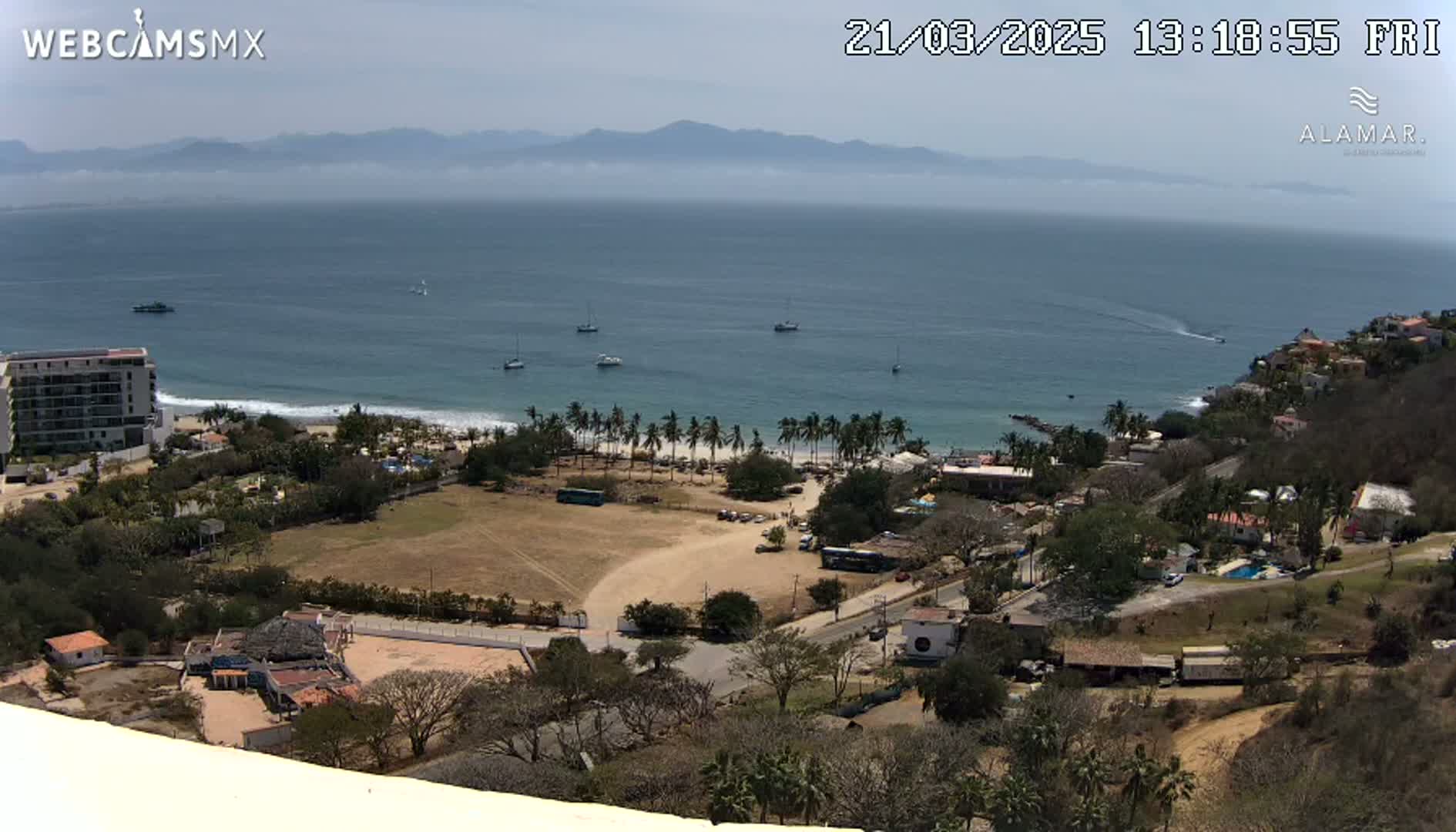 An elevated view of a coastal town on a sunny day, showing a sandy beach with palm trees, buildings, and several boats in the ocean.