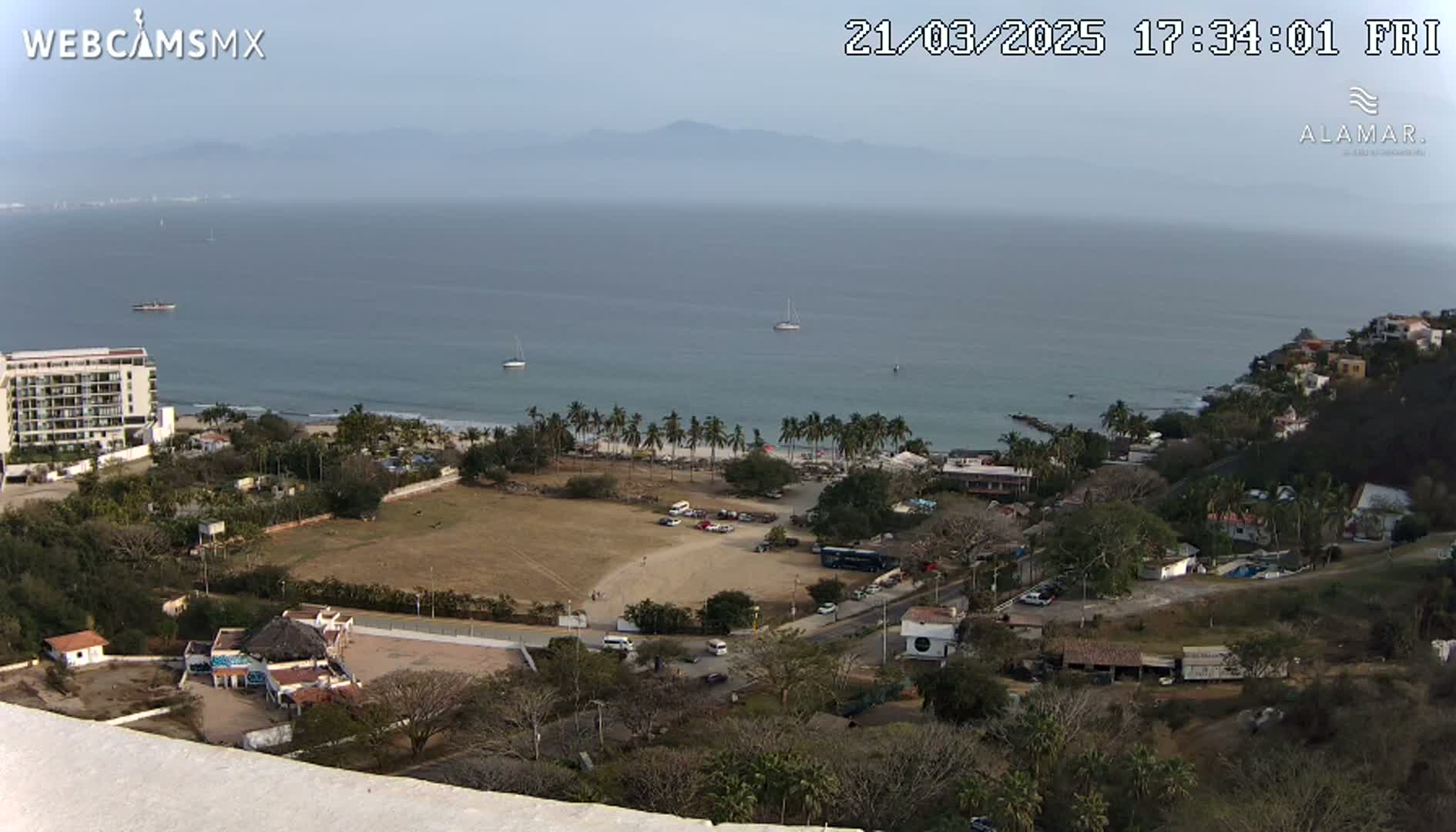 A high-angle view of a coastal town on a partly cloudy day, showing a beach with palm trees, a parking lot, buildings, and a calm ocean with boats in the distance.