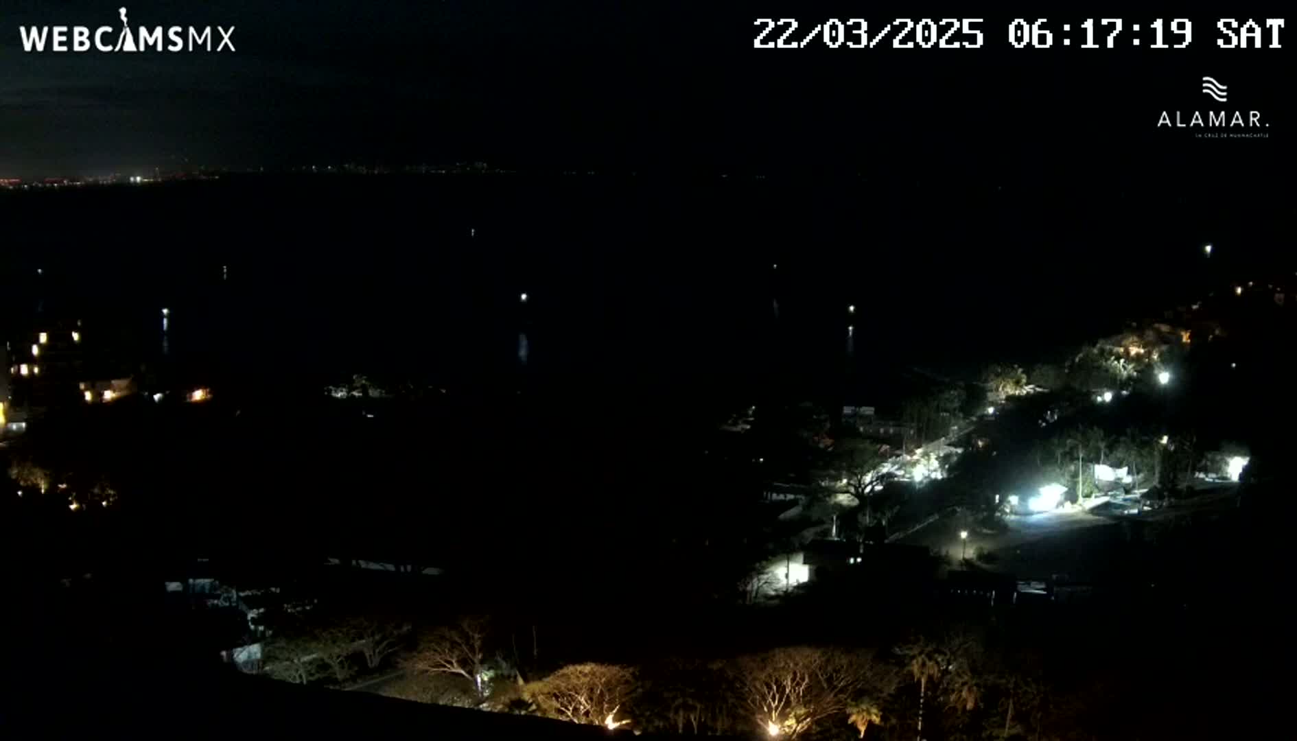A nighttime, high-angle view shows a dark body of water with a few lights visible in the distance and a sparsely lit residential area with trees at the bottom of the frame.