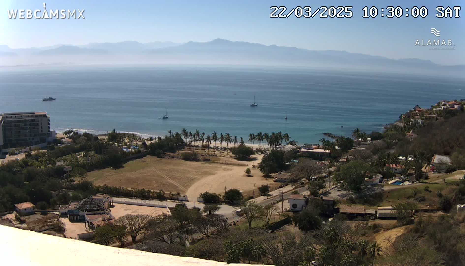An elevated view shows a calm ocean with a few boats, a sandy beach lined with palm trees, coastal buildings, and brown, sparsely vegetated hills under a clear, sunny sky.
