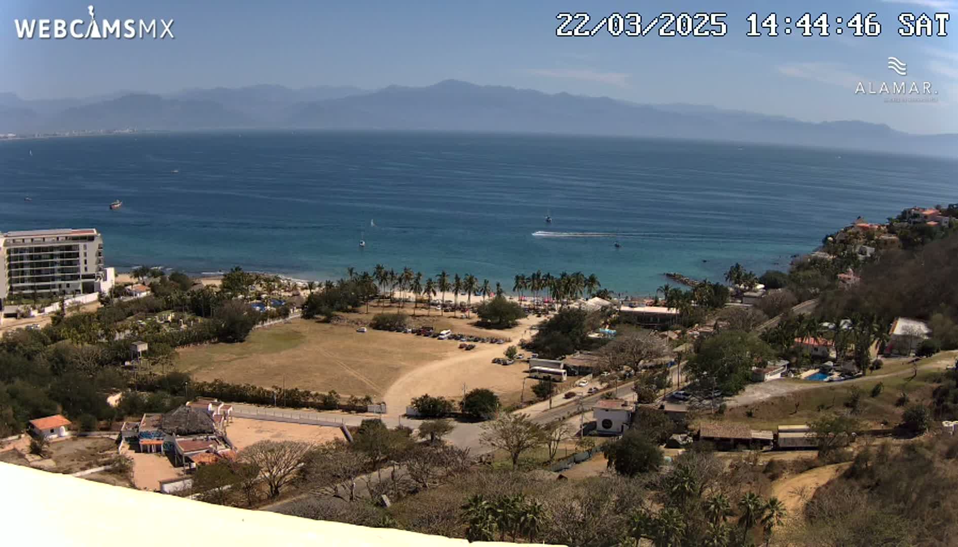 A high-angle view shows a sunny coastal scene with a beach, palm trees, buildings, and a calm ocean under a clear sky, mountains visible in the distance.