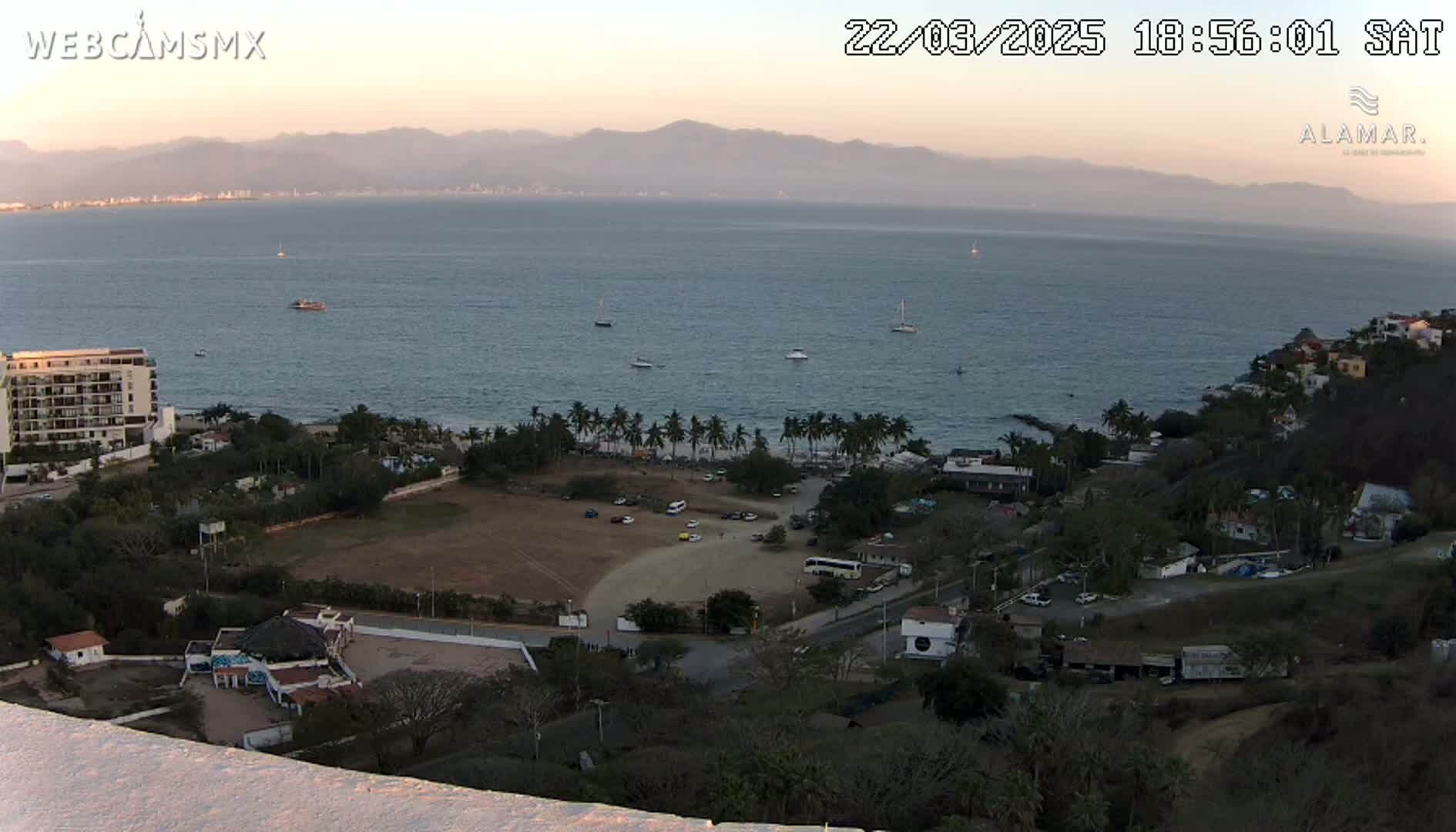 An elevated view of a coastal town at sunset, showing a calm ocean with several boats, a beach with palm trees, and buildings nestled among hills.