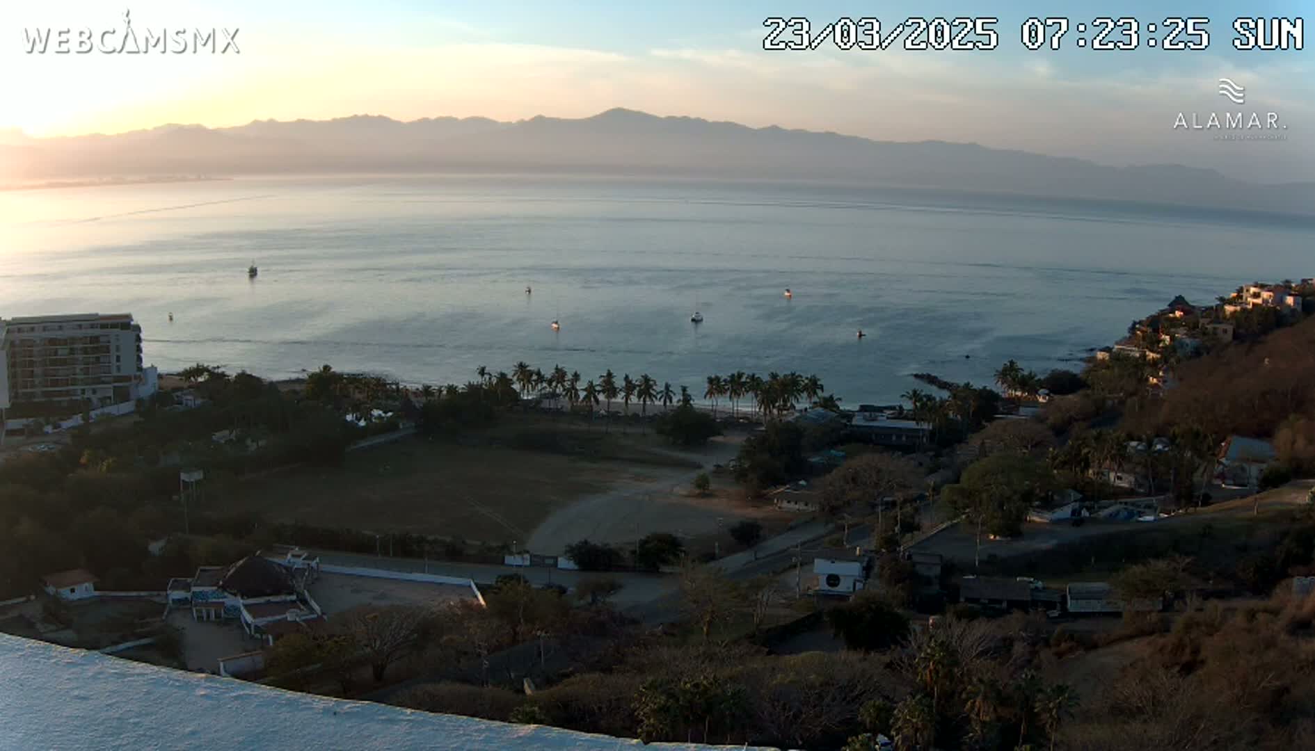 A coastal sunrise view shows a calm ocean with a few boats, a sandy beach lined with palm trees, and a hillside town nestled among brown vegetation, all under a clear sky.