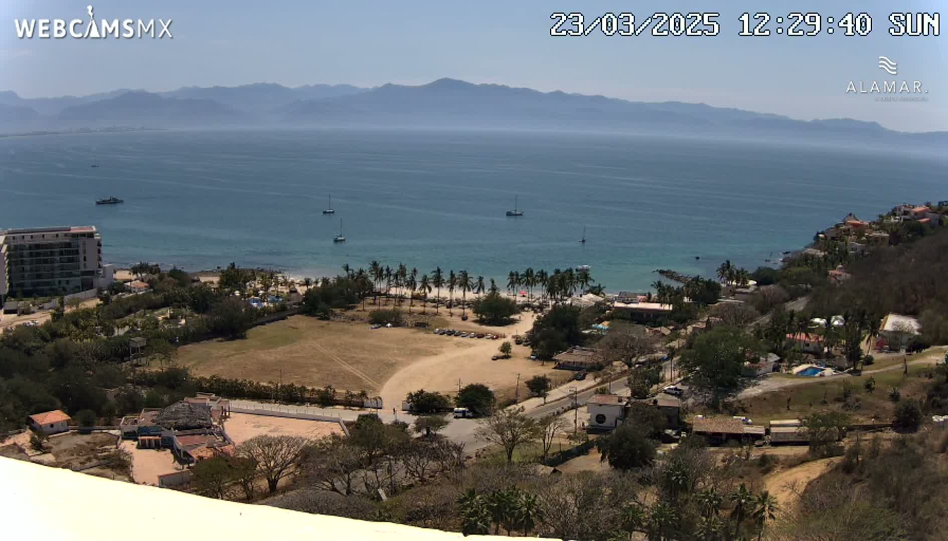 A high-angle, sunny view of a coastal town with a sandy beach, palm trees, buildings, and a calm ocean with mountains in the distance.