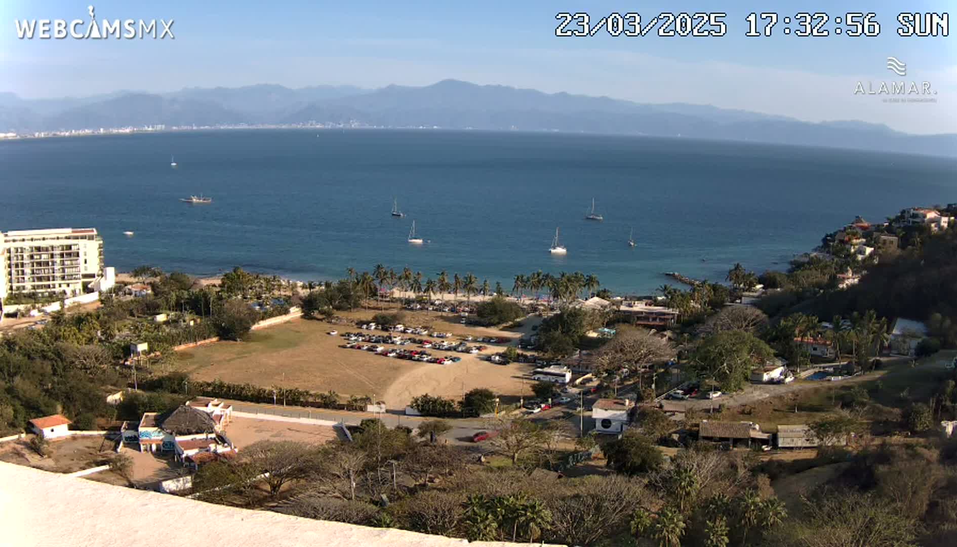 A sunny, high-angle view shows a coastal town with a parking lot, beachfront, and several sailboats on a calm bay with mountains in the background.