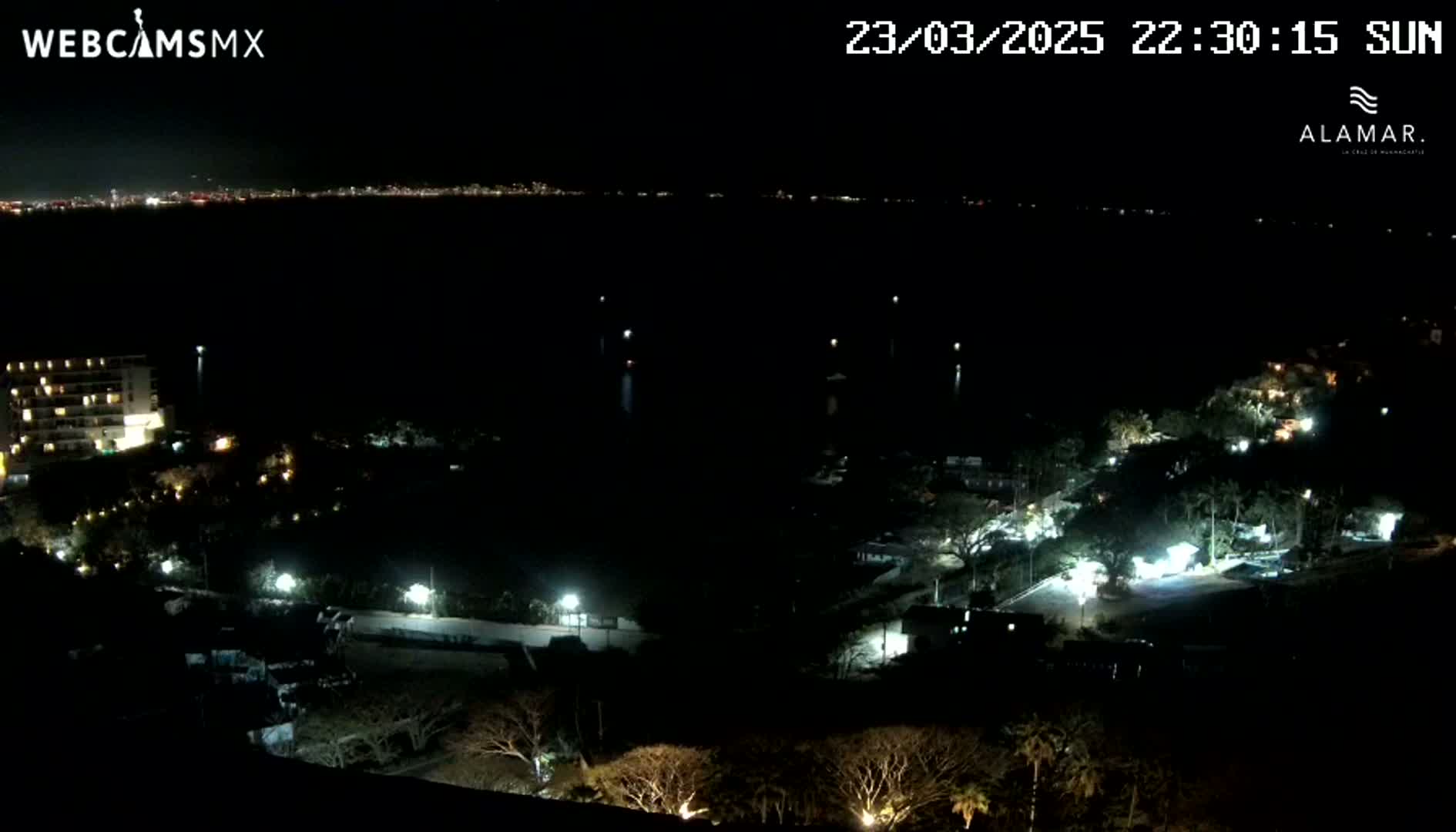 A nighttime view shows a dark body of water with a distant city skyline, and a land area with sparsely lit buildings and trees.