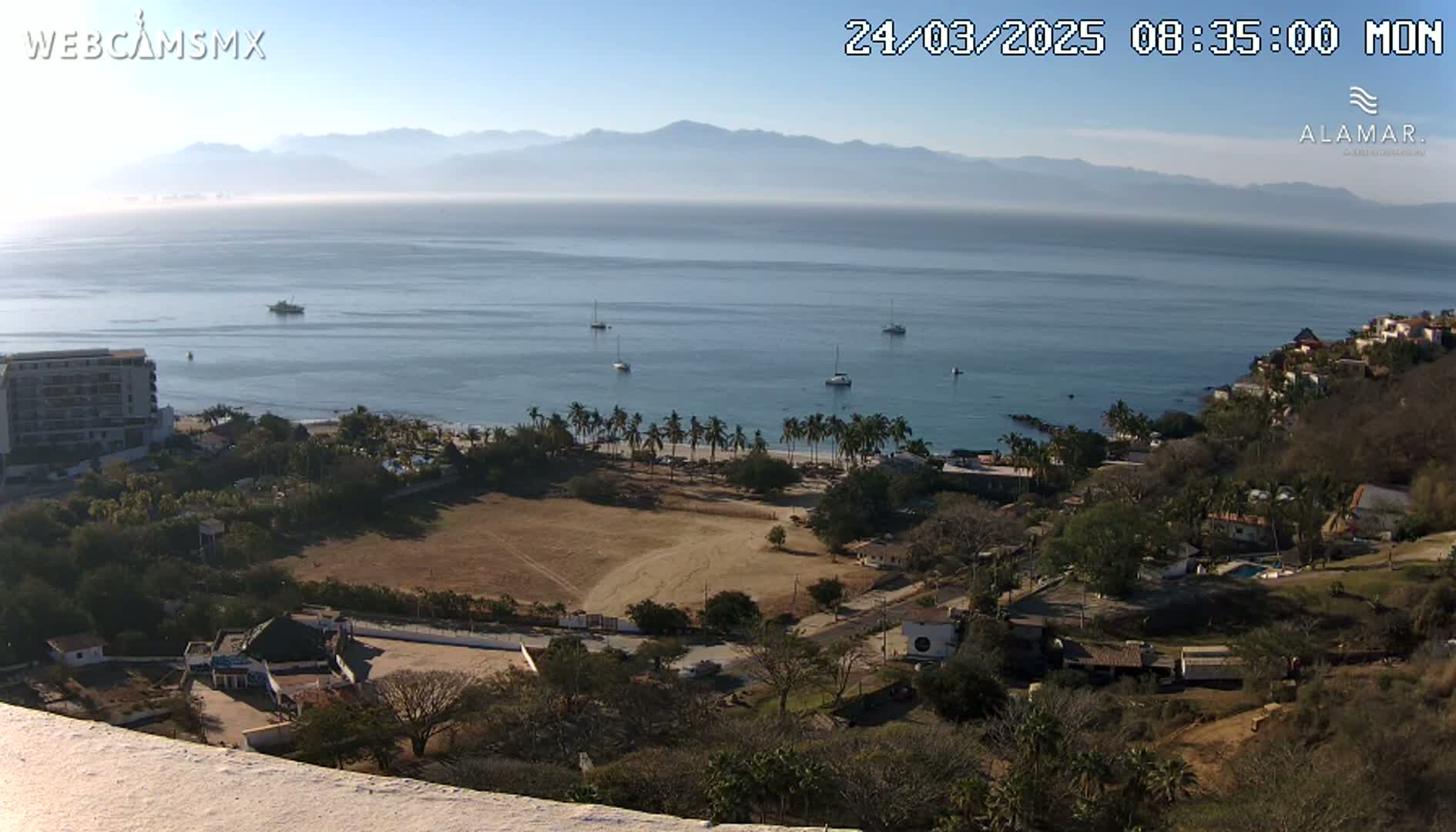 A high-angle, long shot of a coastal town on a sunny day, with a calm ocean, several boats, and a hazy mountain range in the distance.