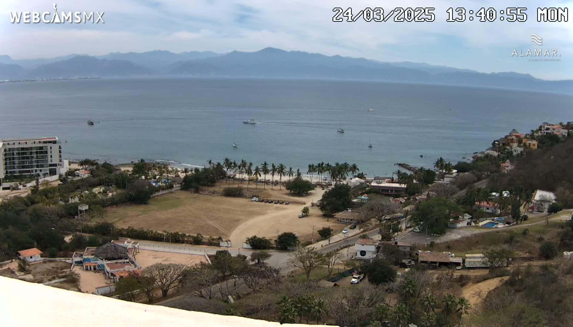 An elevated view of a coastal town on a partly cloudy day, showing a sandy beach with palm trees, a calm ocean with boats, and mountains in the distance.