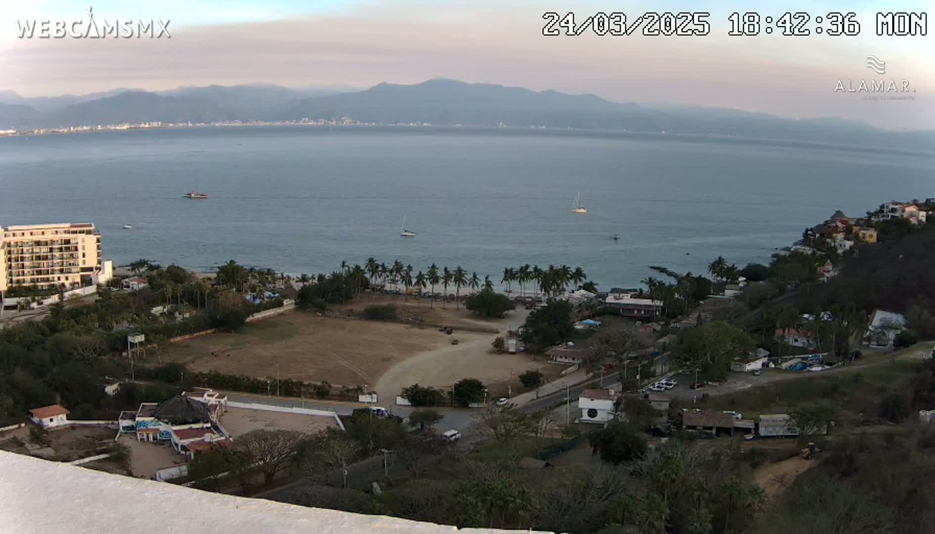 An elevated view of a coastal town on a partly cloudy day, showing a calm bay with boats, a sandy beach with palm trees, buildings, and dry, brown land.