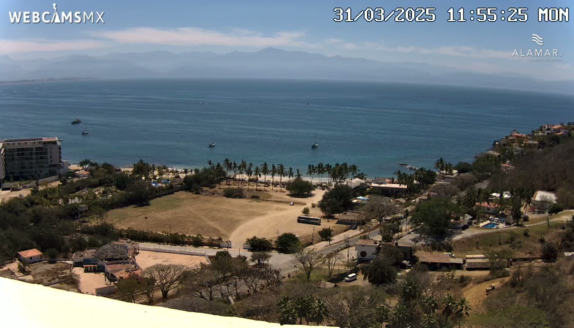 A high-angle, sunny view of a coastal town with a sandy beach, palm trees, and a calm ocean extending to distant mountains under a mostly clear sky.