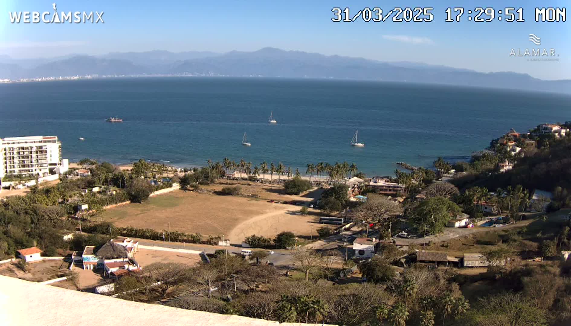 An elevated view of a coastline on a sunny day shows a calm ocean with several sailboats, a sandy beach lined with palm trees, buildings, and vegetation extending into the background where mountains are visible.
