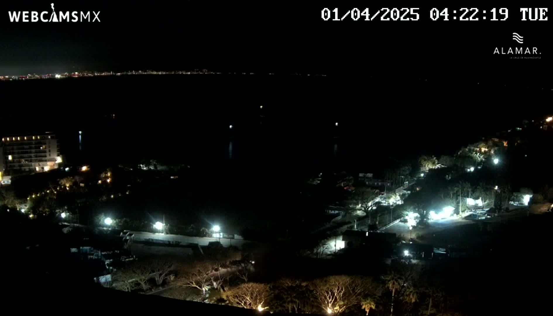 A nighttime view shows a dark body of water with a city skyline in the distance, and a sparsely lit area of land with buildings and trees in the foreground.