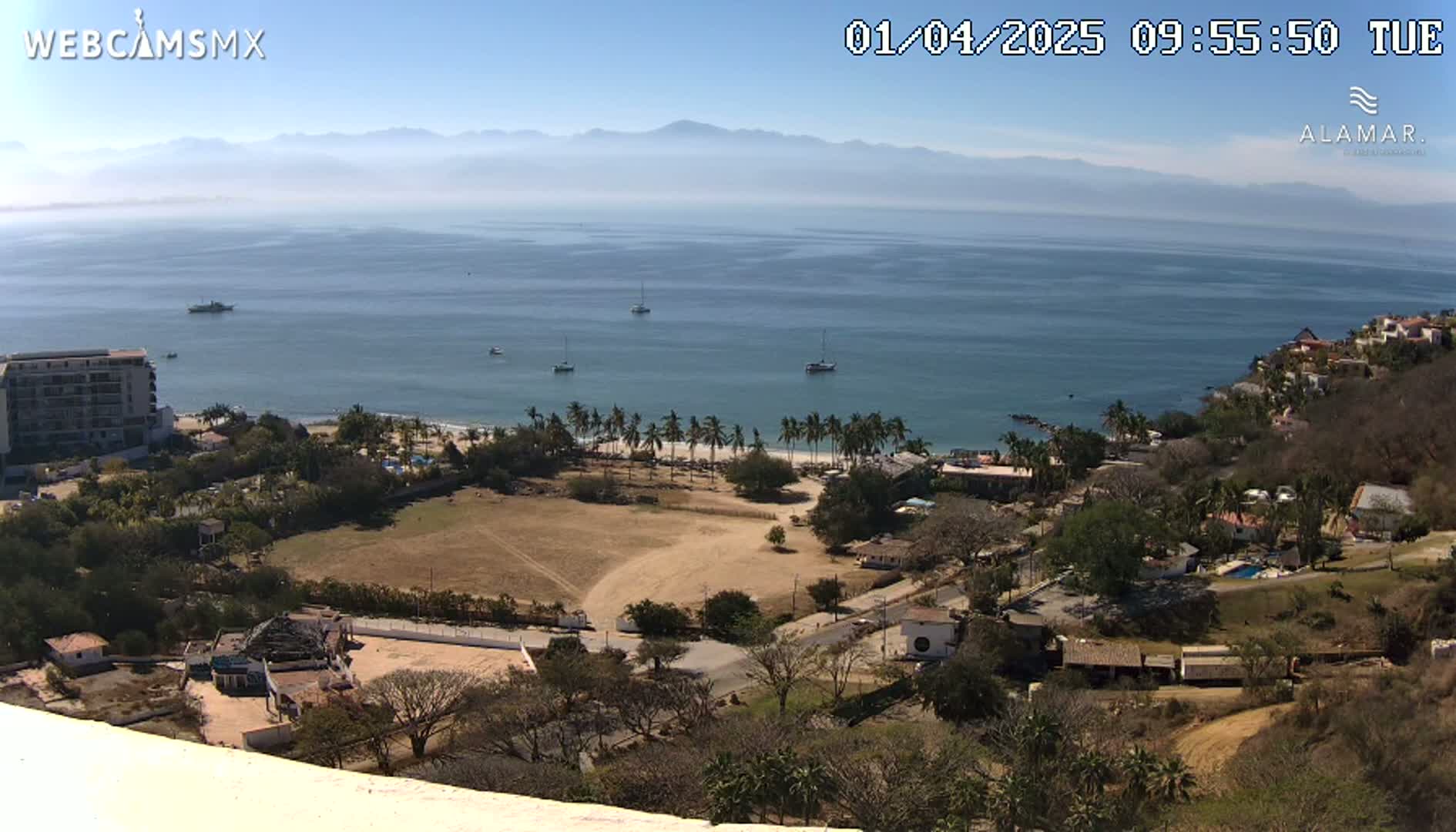 A high-angle, long shot on a clear, sunny day shows a sandy beach with palm trees, a calm ocean with several boats, and a coastal town nestled against a hillside, all with mountains faintly visible in the distance.