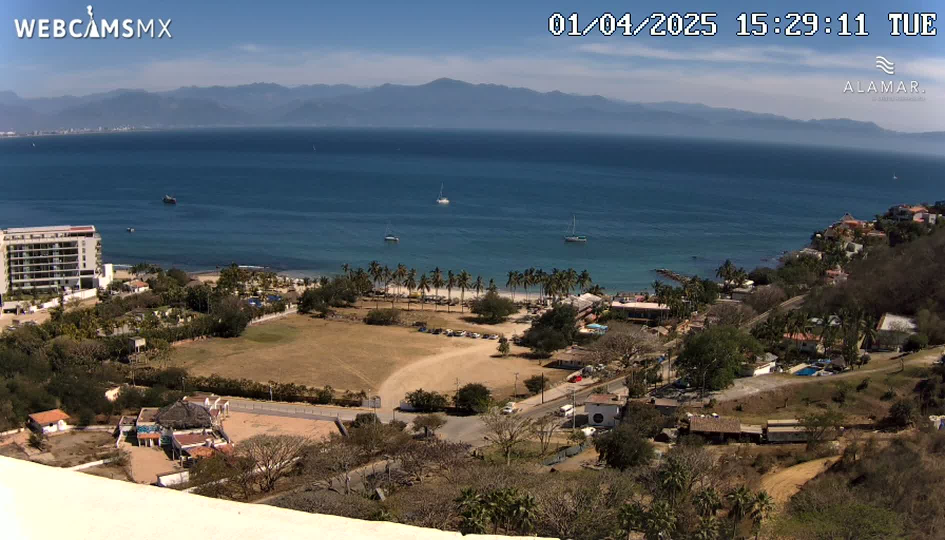 A high-angle, sunny view shows a coastline with a sandy beach, palm trees, buildings, and a calm ocean with a few boats under clear skies and mountains in the distance.