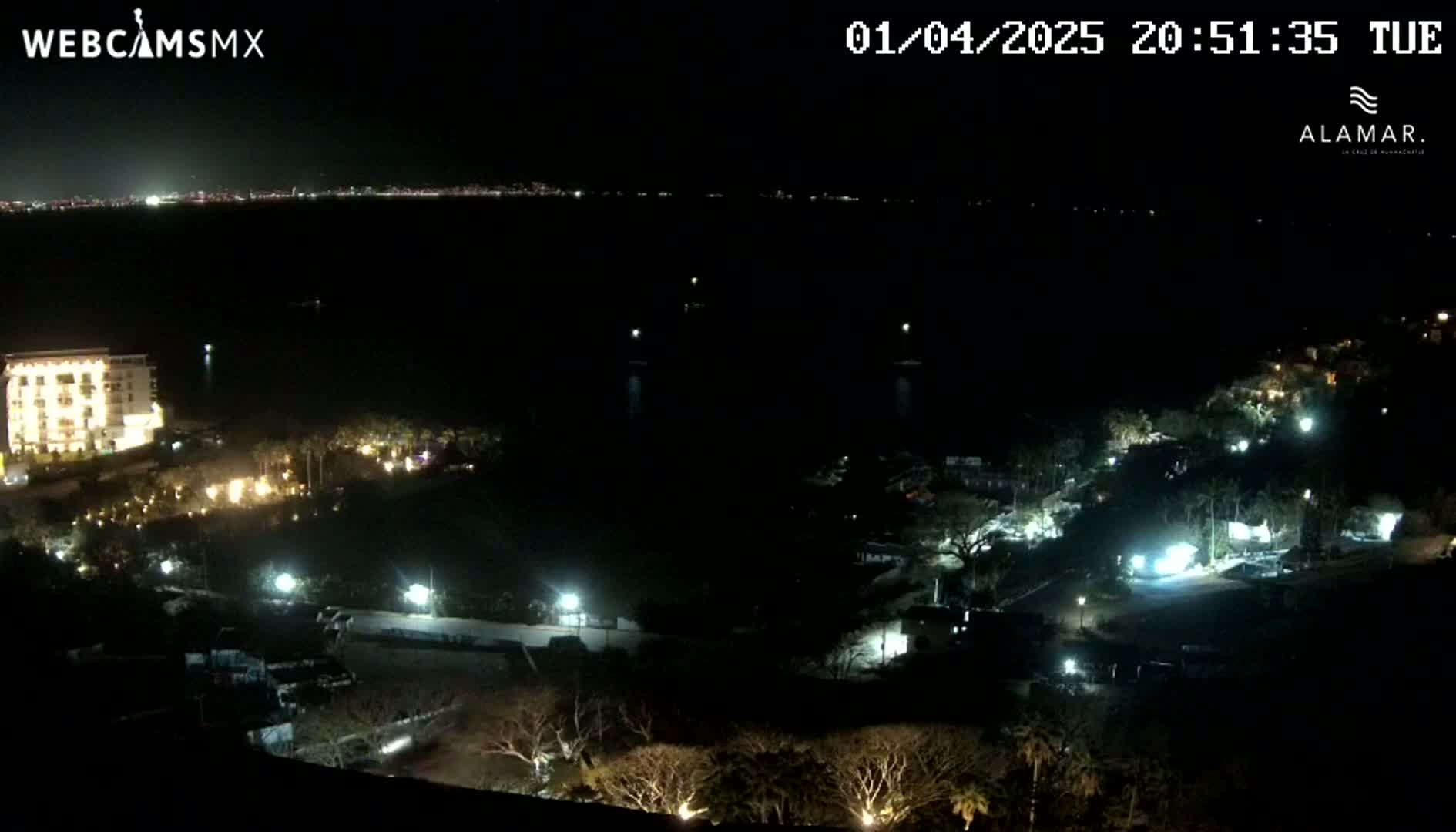 A nighttime view of a coastal area shows a body of water, a brightly lit hotel, and other buildings with lights on, under a clear, dark sky.