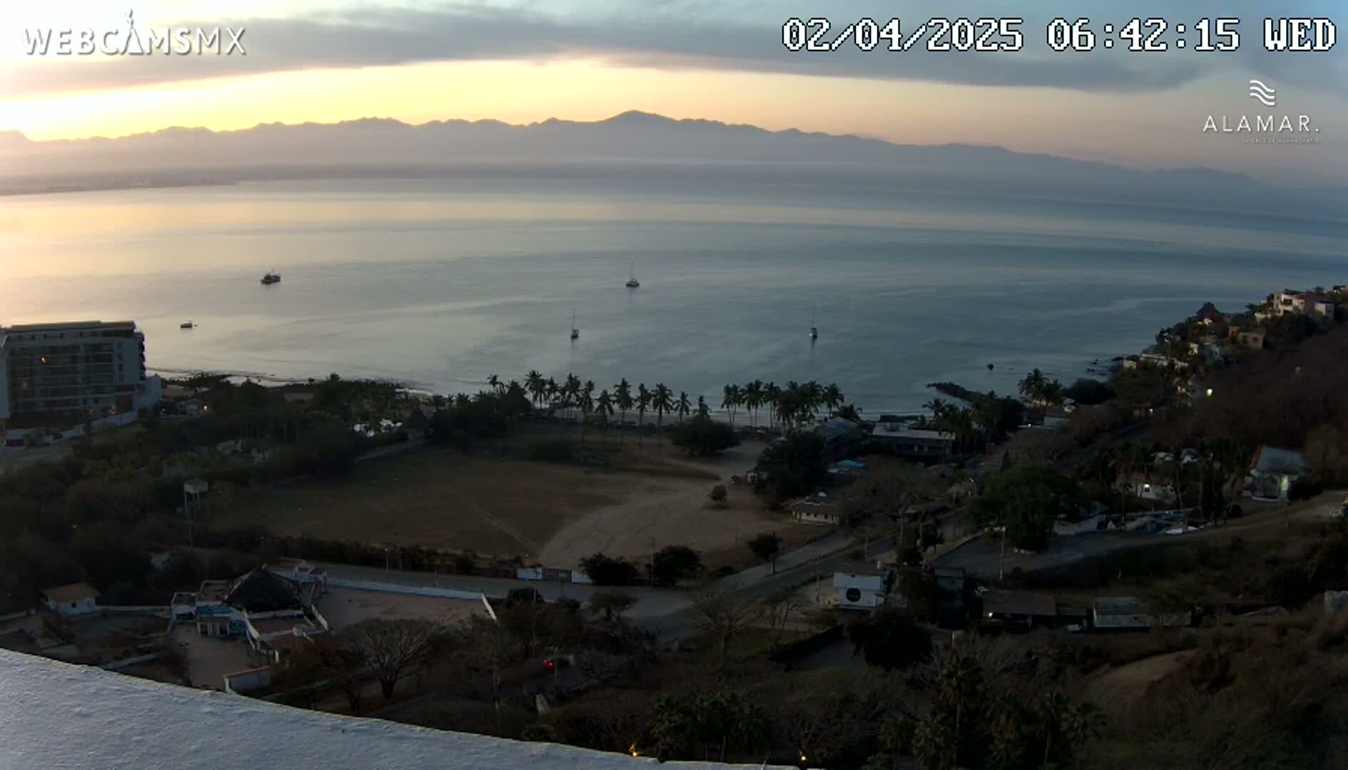 A high-angle, long shot of a calm ocean at sunrise, with a coastline featuring buildings, palm trees, and a sandy area, all under a mostly clear sky.