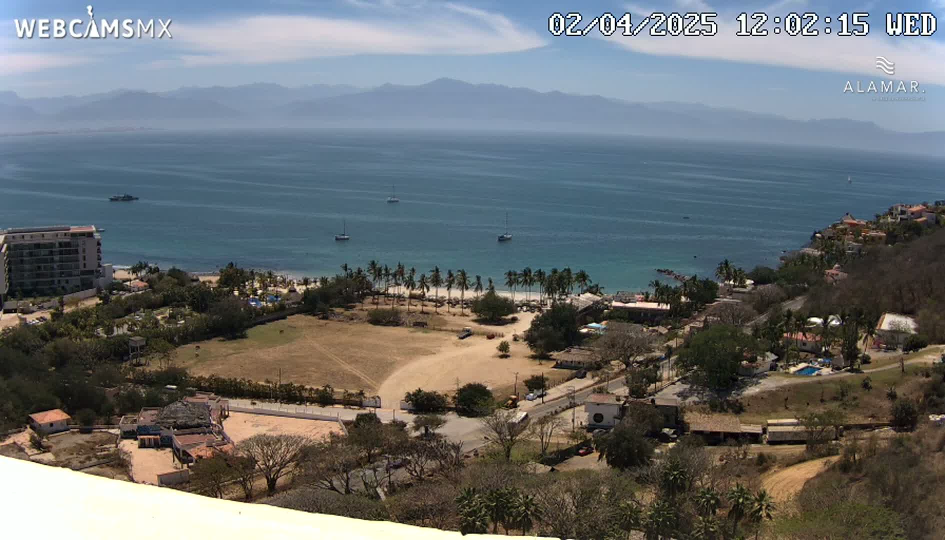 An elevated view of a coastal town on a sunny day, with a calm ocean extending to distant mountains and several boats visible on the water.