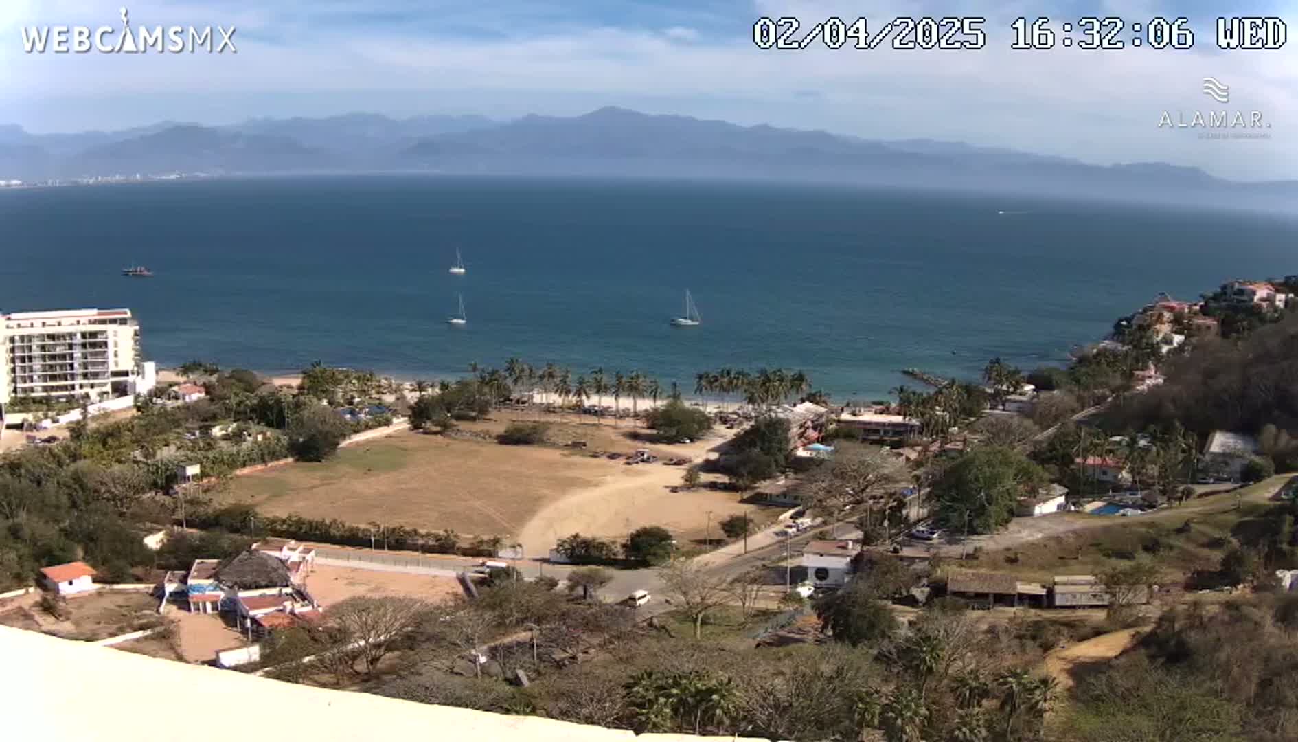 An elevated view of a coastal town on a sunny day, showcasing a calm ocean with several sailboats, a sandy beach lined with palm trees, and a mix of buildings and dry, brown land.