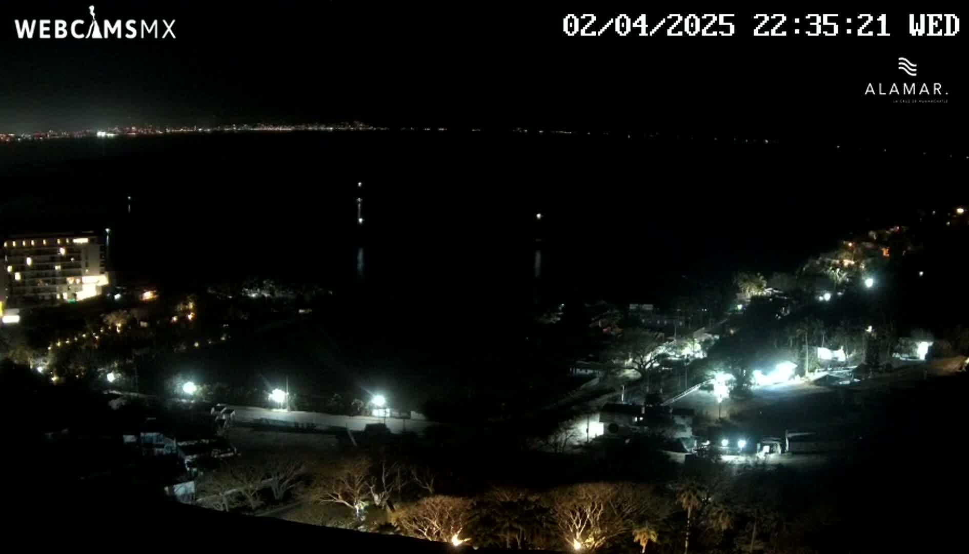 A nighttime view of a coastal area shows a dark body of water with faintly lit distant city lights, a dark landscape with scattered lights from buildings and trees, and a multi-story building near the water's edge.