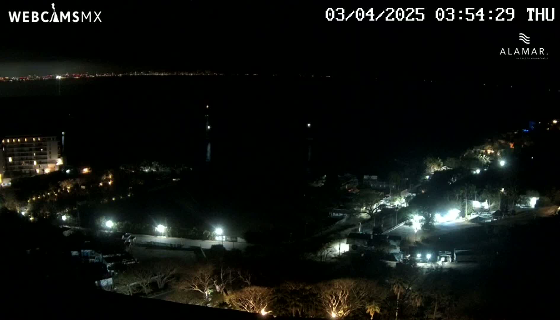 A nighttime, aerial view shows a coastal area with sparsely lit buildings and vegetation, overlooking a dark body of water with a distant city skyline visible on the horizon.