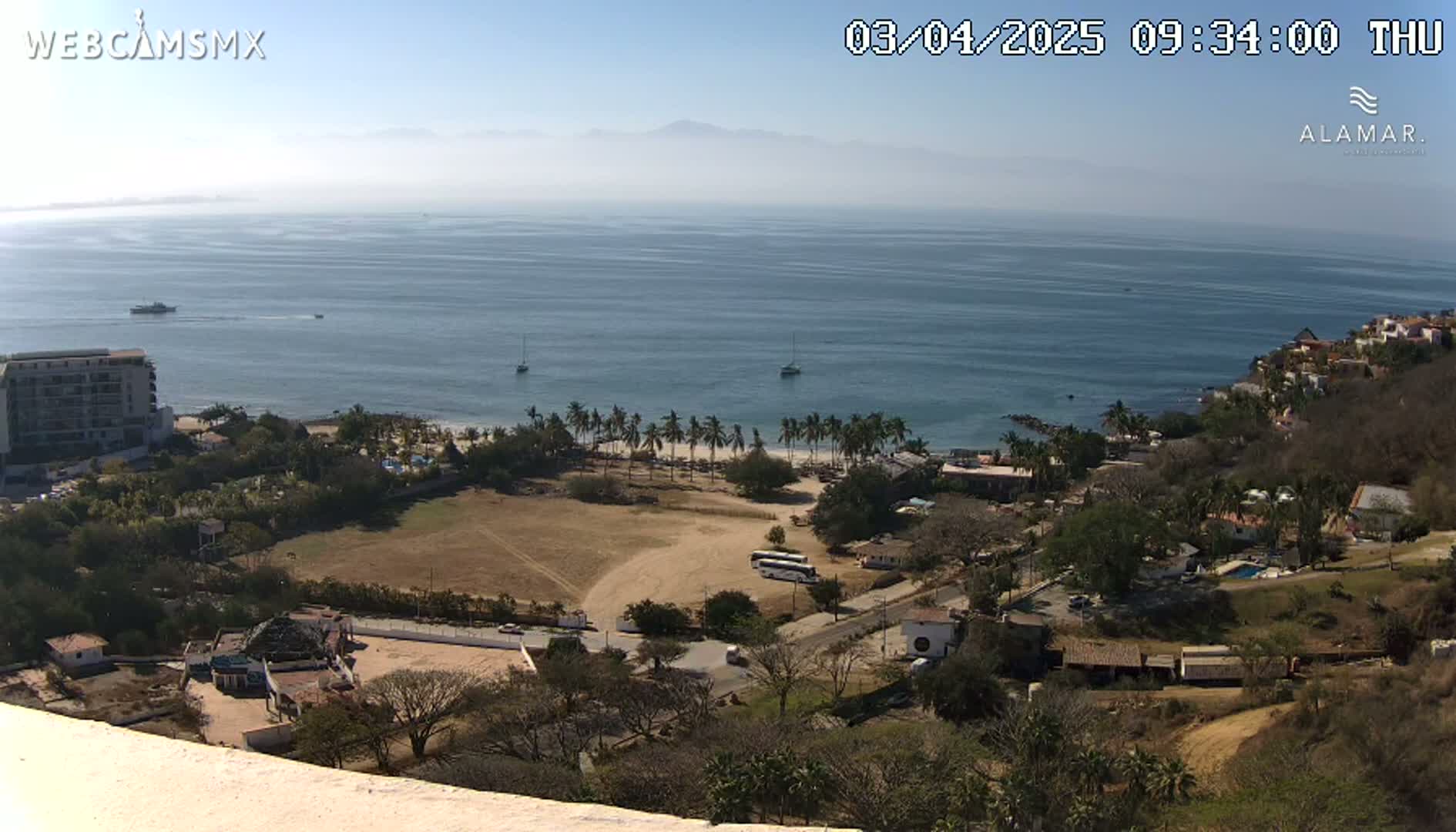 An elevated view of a coastal town on a sunny day, showing a sandy beach, palm trees, buildings, and a calm ocean extending to distant mountains.