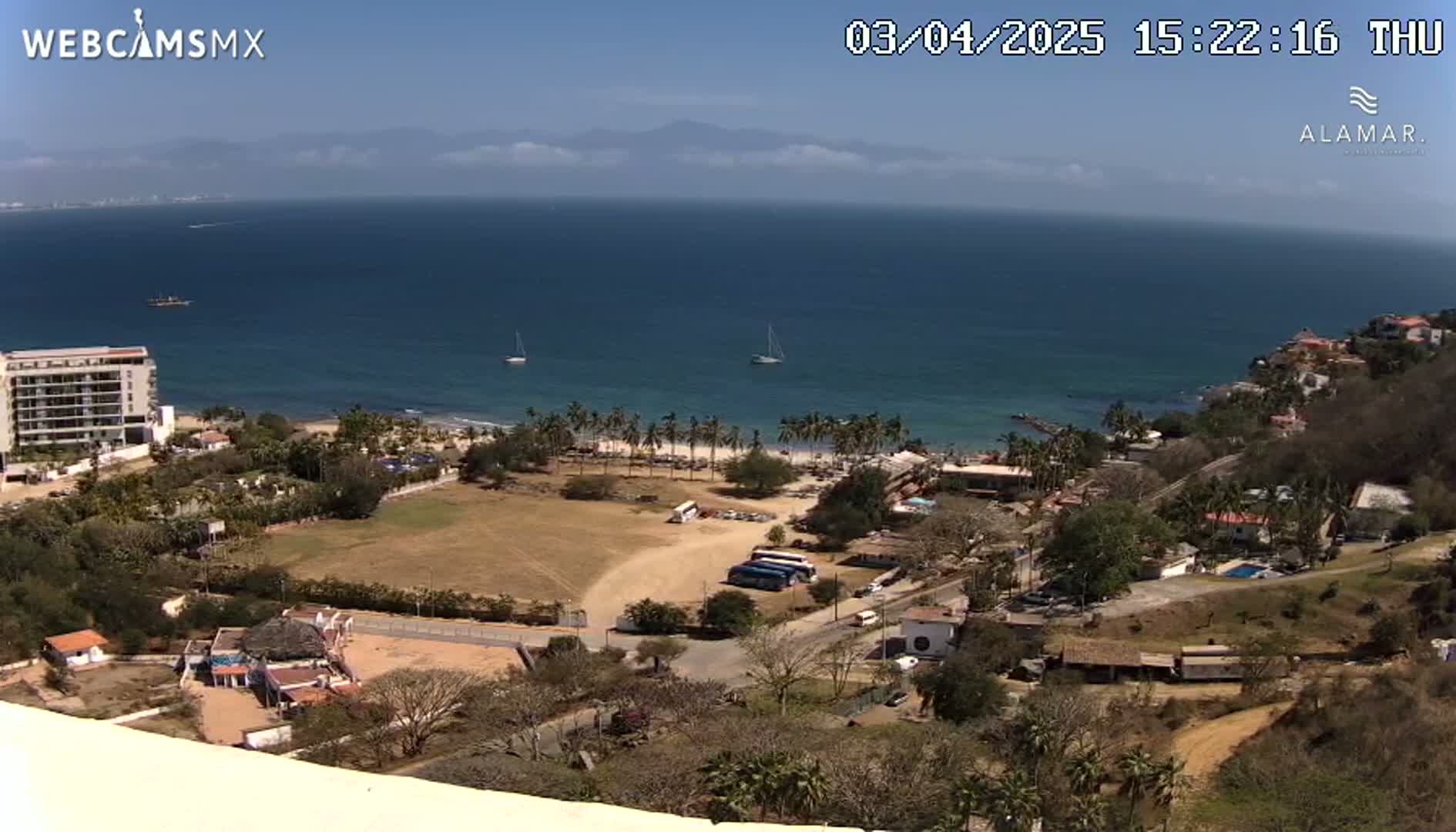 An elevated view of a coastal town on a sunny day shows a calm ocean, a sandy beach lined with palm trees, and buildings nestled among sparse vegetation.