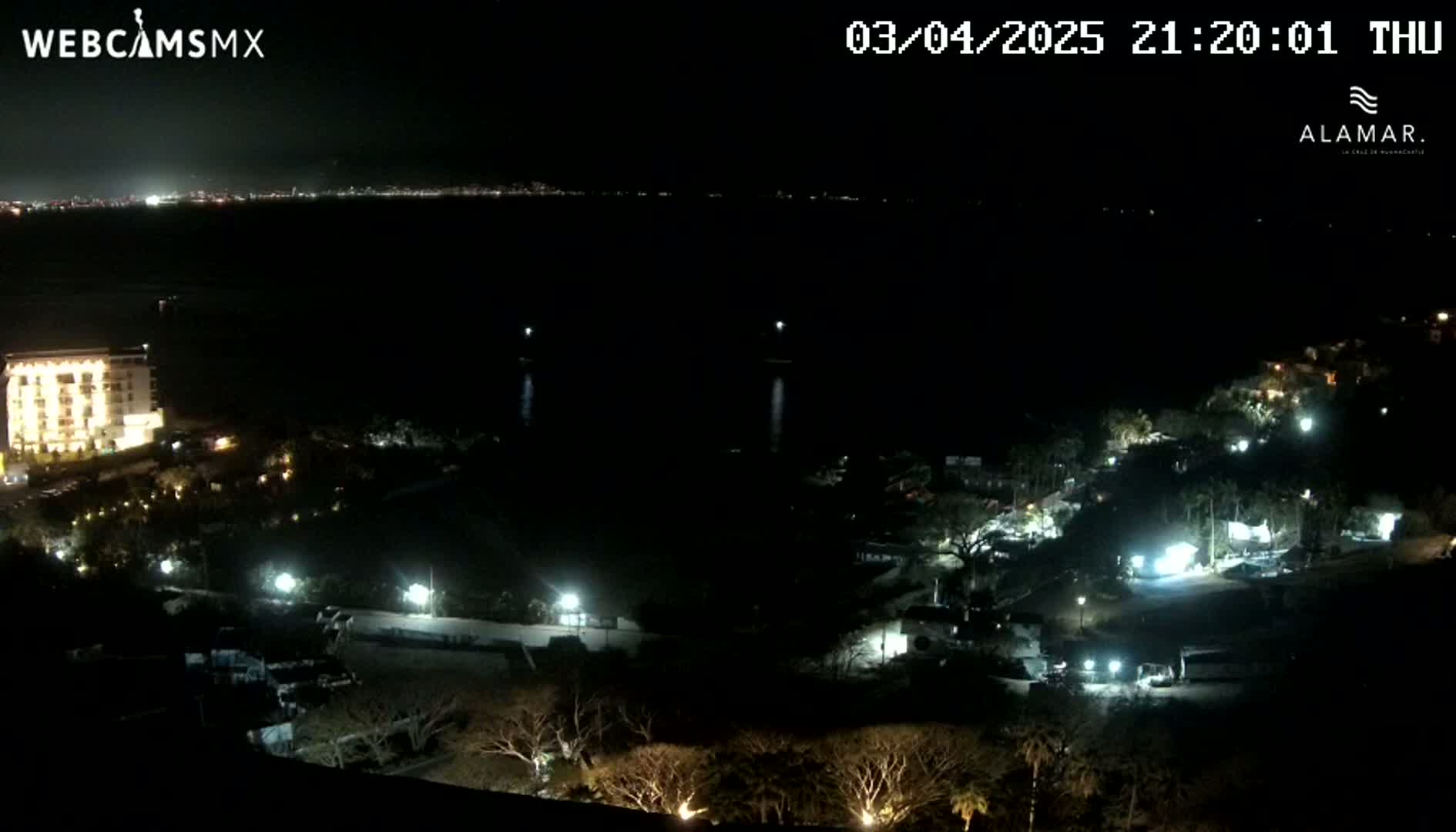 A nighttime, high-angle view of a coastal area shows a resort with illuminated buildings and landscaping, and a dark body of water with a distant city skyline faintly visible in the background.