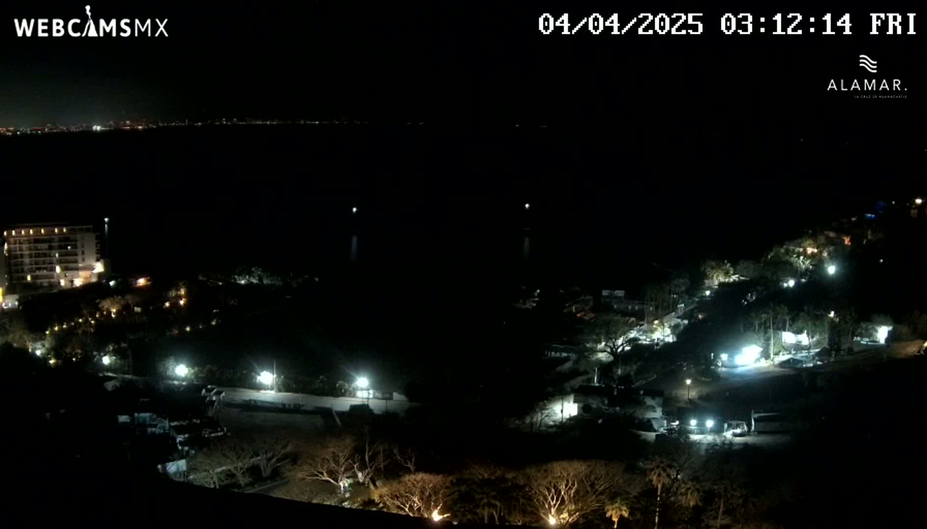 A nighttime view of a coastal area shows a dark body of water, a lit-up building, and sparsely lit landscaping with trees.