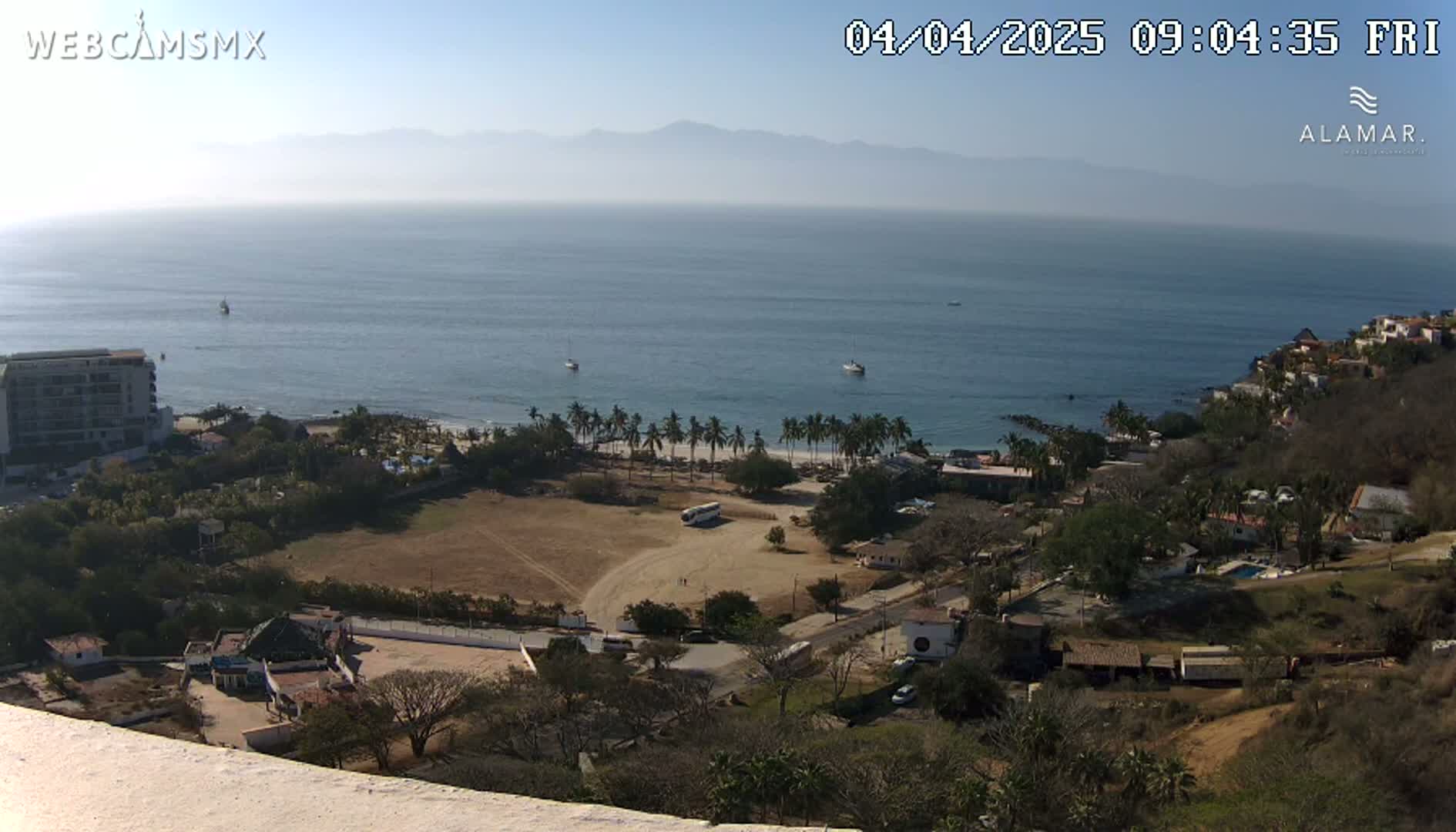 A high-angle view of a coastal town on a sunny day, showing a sandy beach, palm trees, buildings, and a calm ocean with hazy mountains in the distance.