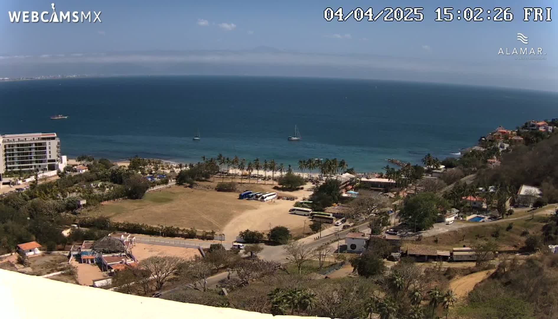 A high-angle, sunny view shows a beach with palm trees, several buildings, a parking lot with buses, and a calm ocean under a clear sky.