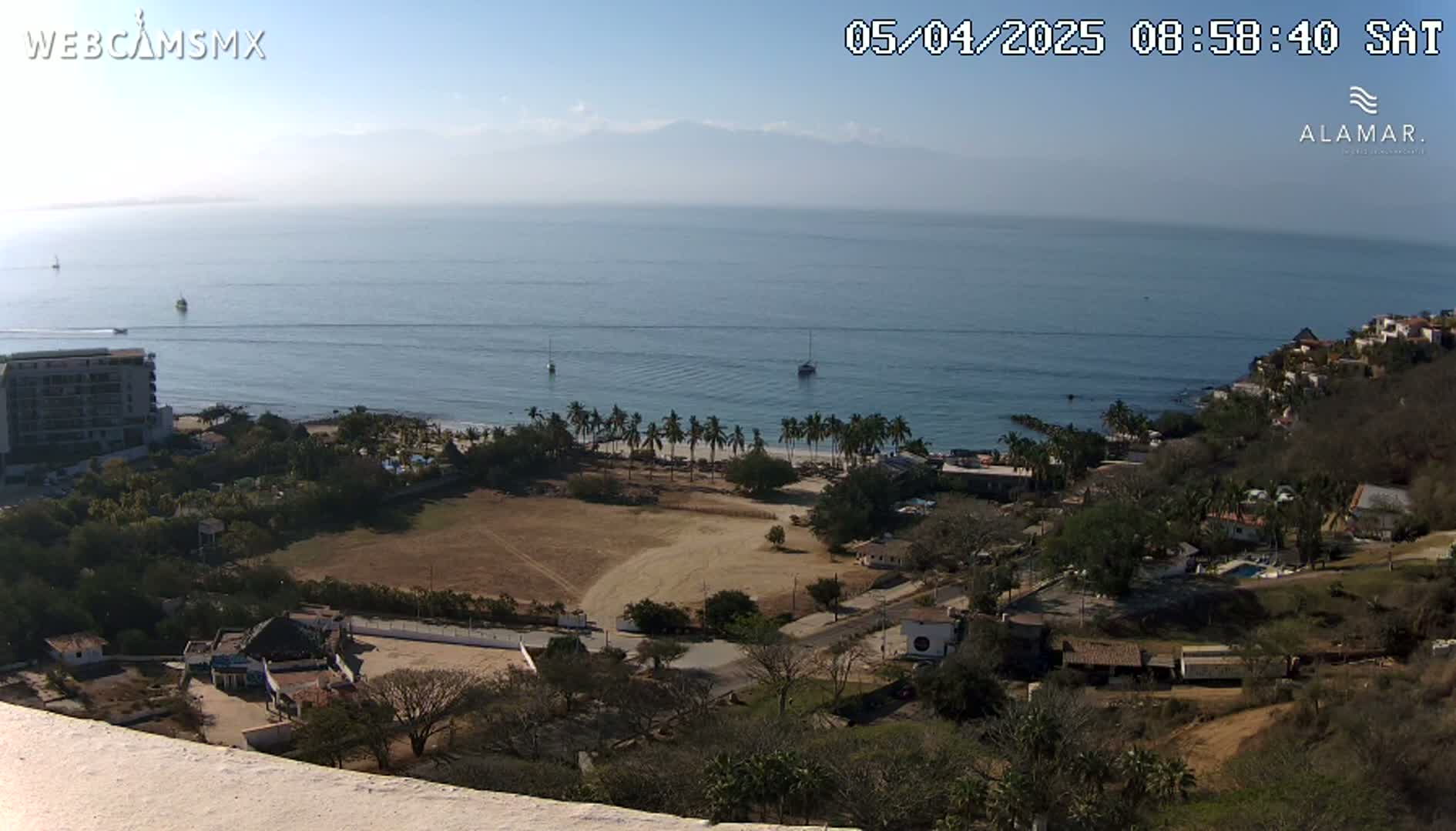 A high-angle, long shot on a clear, sunny day shows a coastline with a sandy beach, palm trees, buildings, and a large, empty field, all overlooking a calm ocean with a few boats.