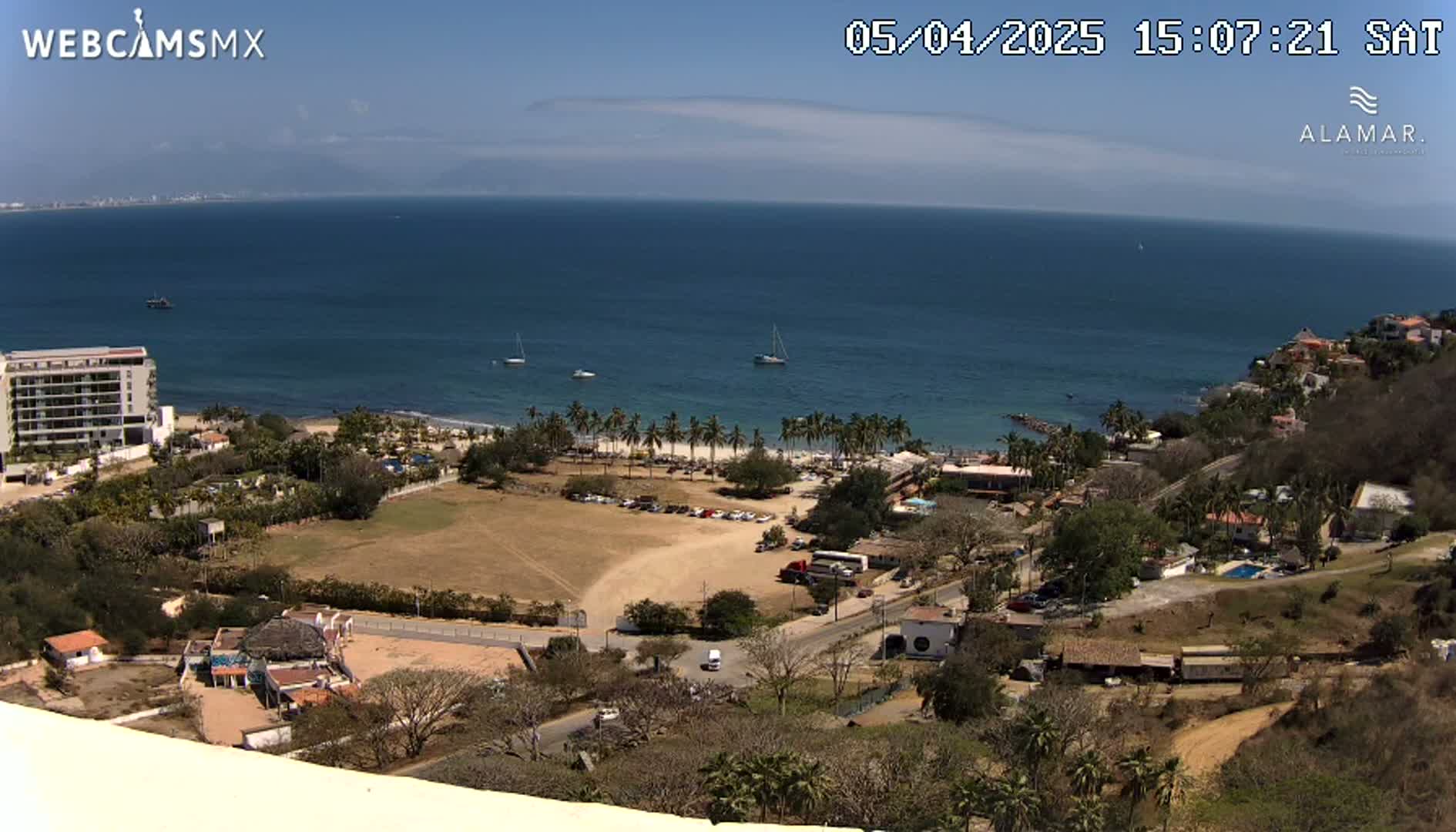 An aerial view of a coastal town on a sunny day, showing a sandy beach, palm trees, buildings, and a calm ocean with a few boats.